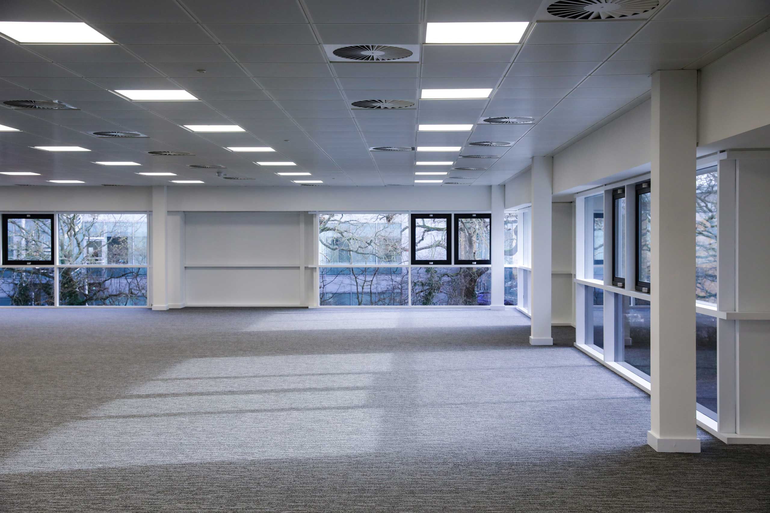 Empty modern office space with large windows, gray carpet, and ceiling lights.