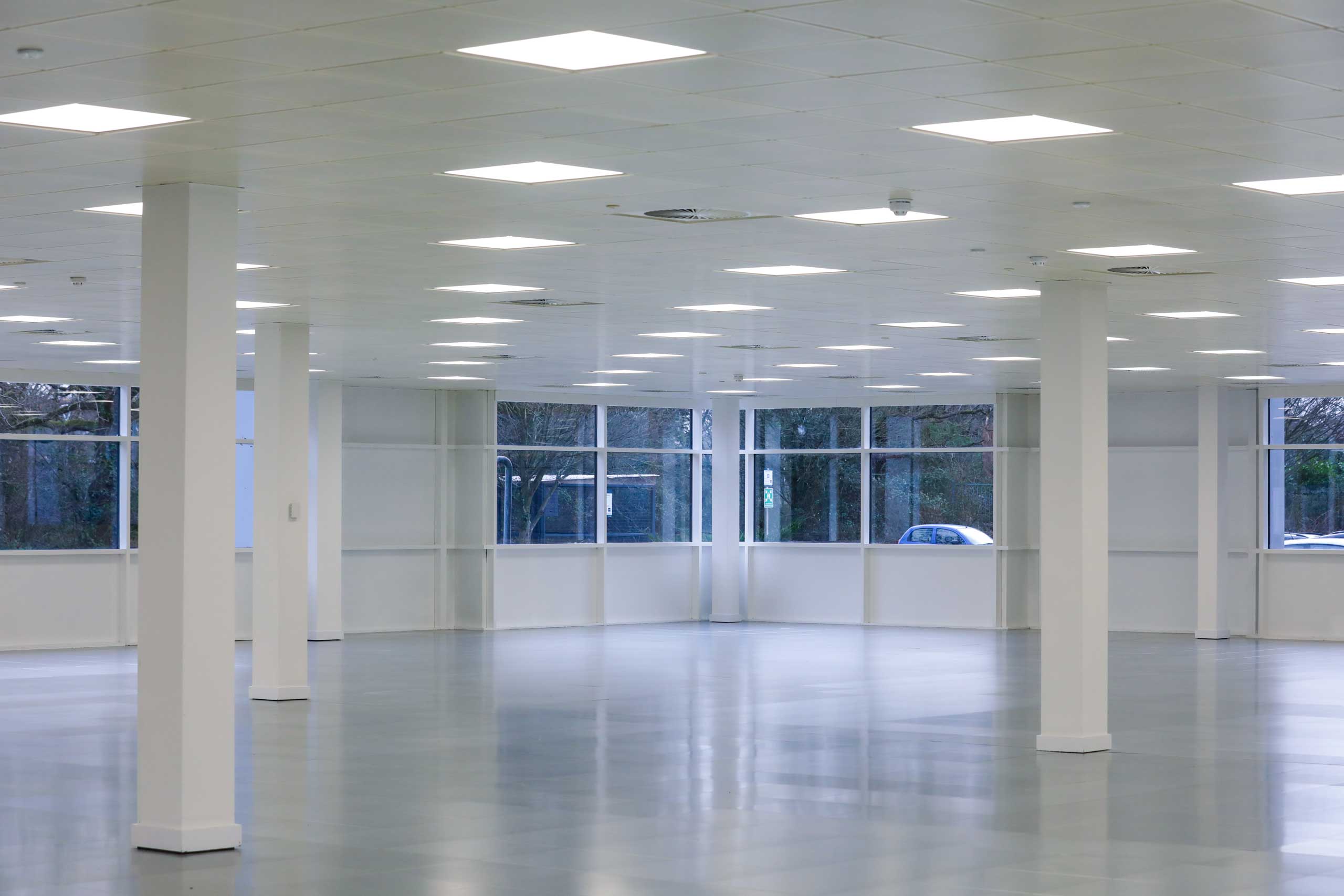 Spacious, empty office with white walls, large windows, and ceiling lights turned on.