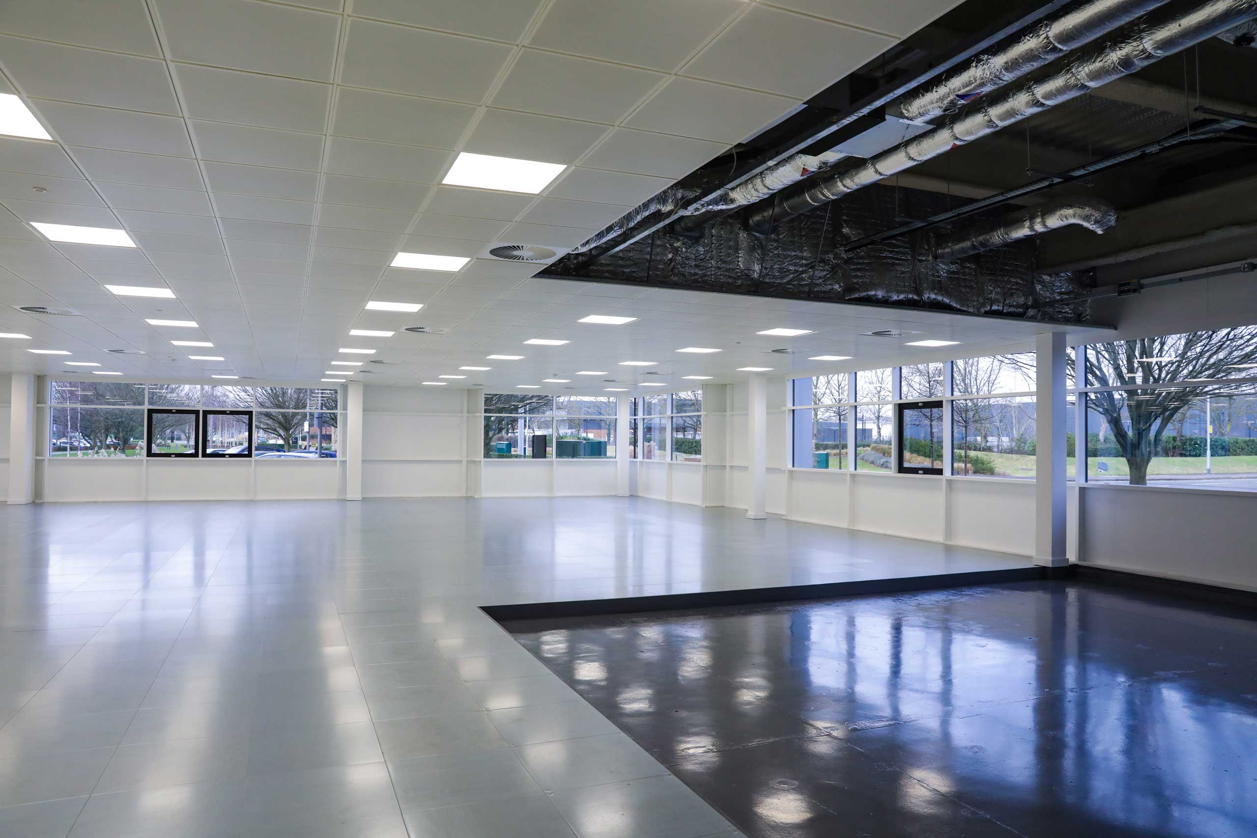 Large empty room with tiled floor, bright ceiling lights, and exposed ductwork near the windows.