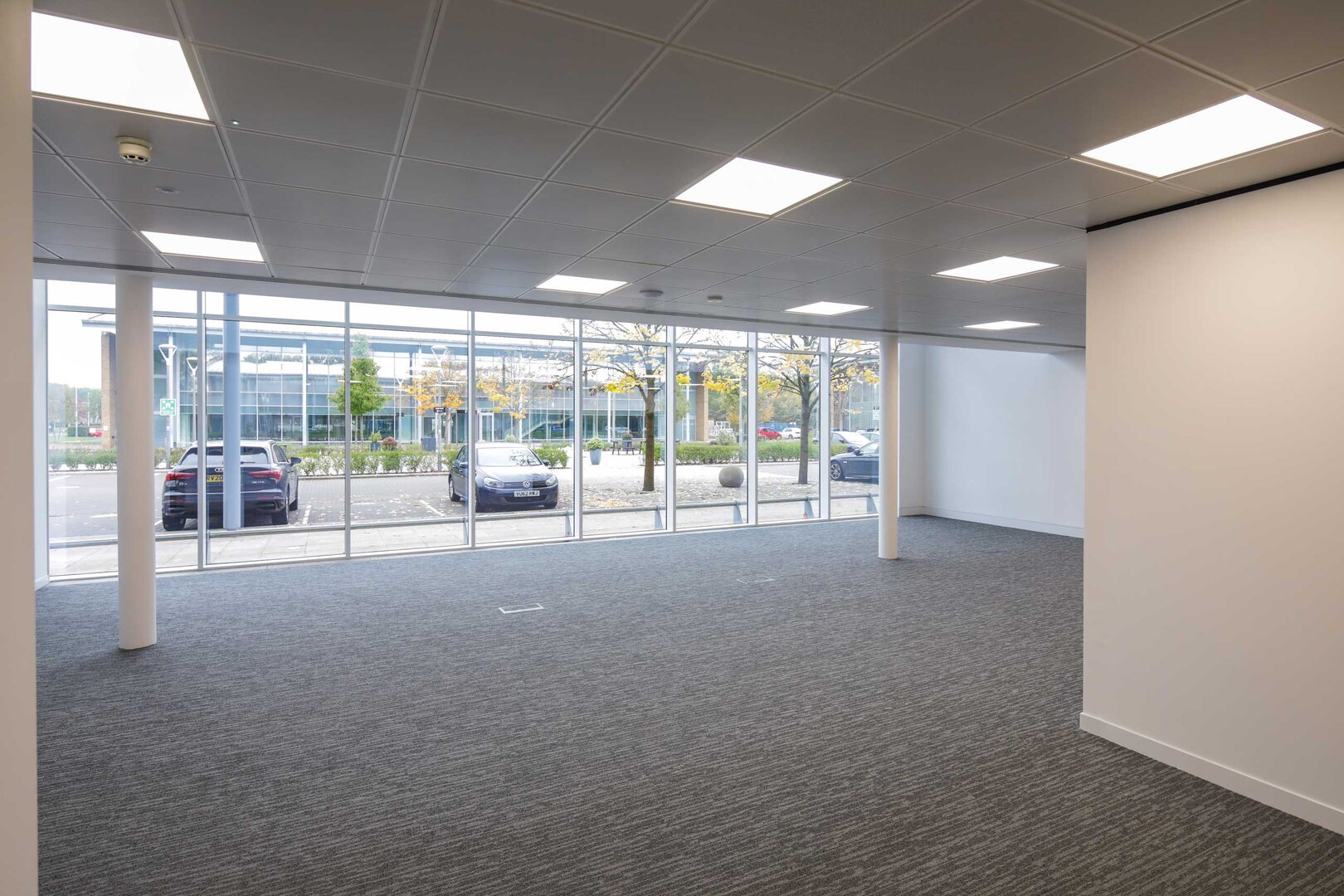 Empty office space with carpet flooring, large windows, and ceiling lights, facing a parking lot.
