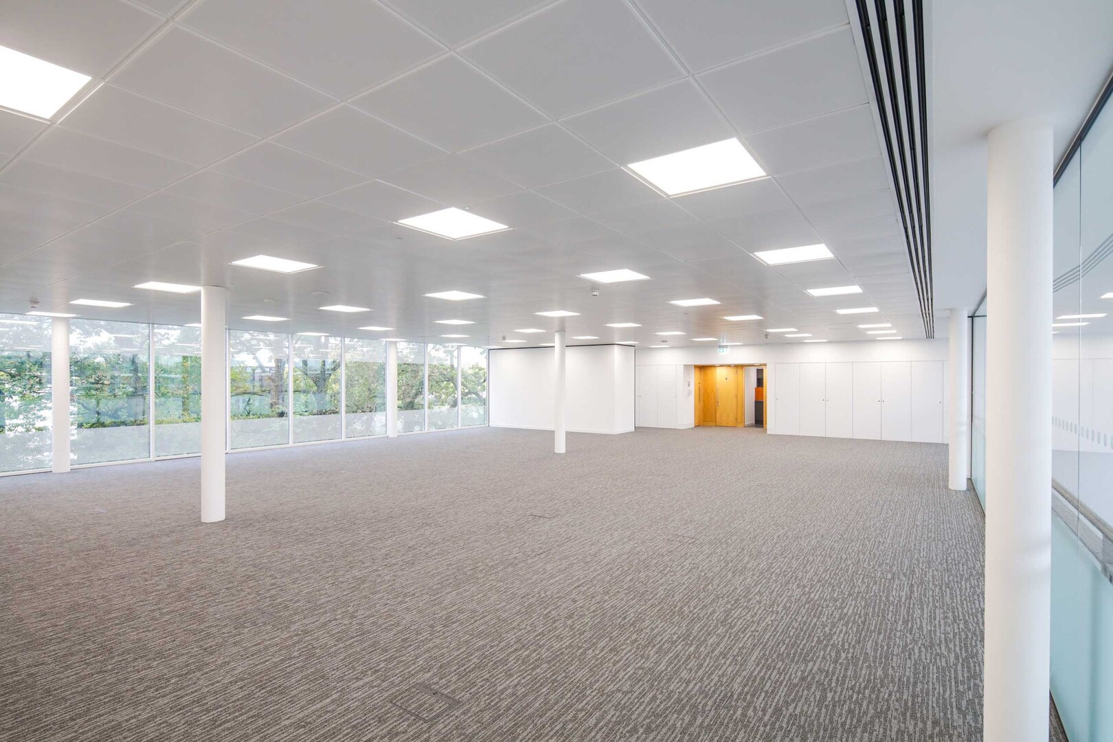 Spacious, empty office room with carpeted floor, large windows, white walls, and ceiling lights.