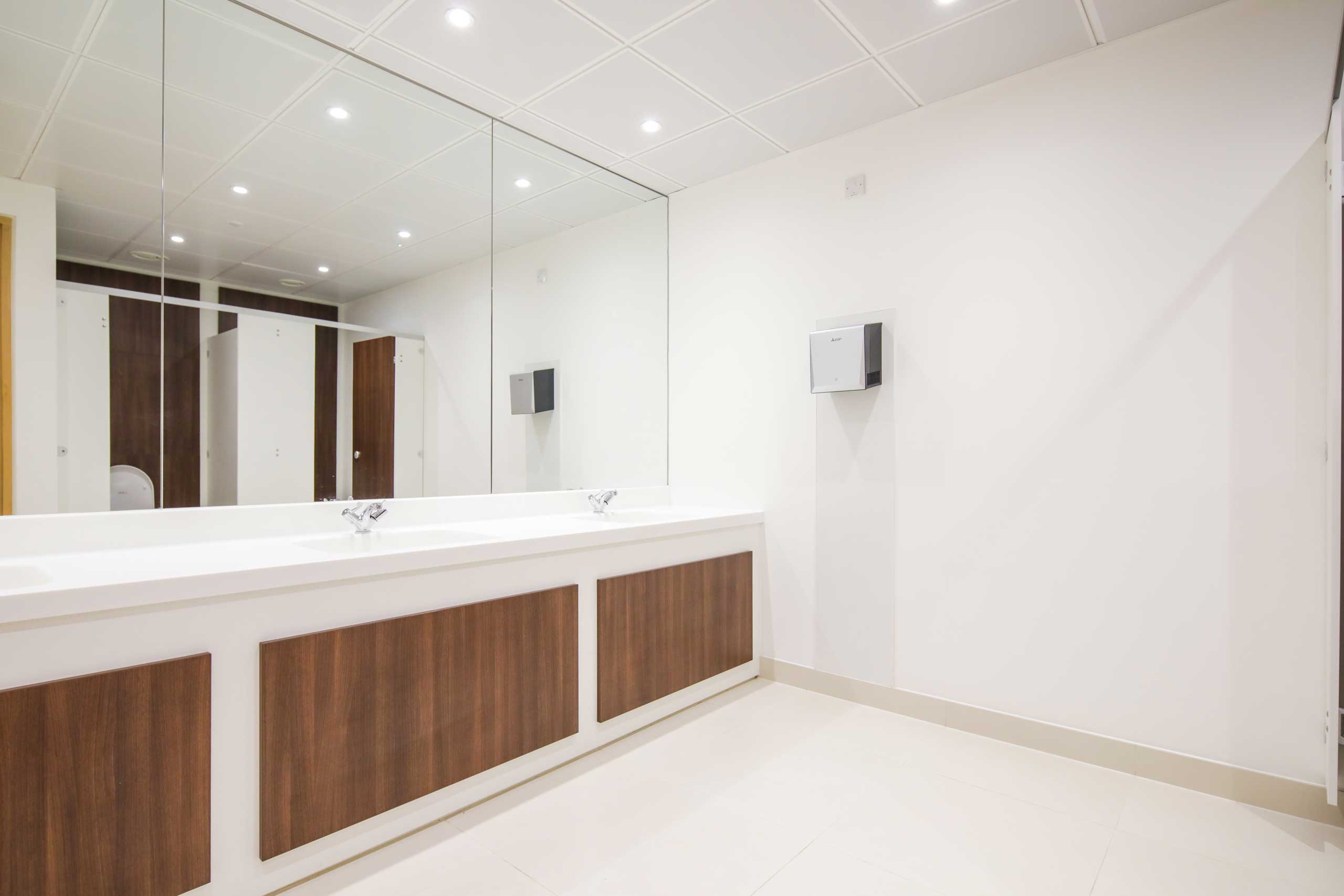 Modern public restroom with large mirrors, white sinks, wood panel accents, and toilet stalls.