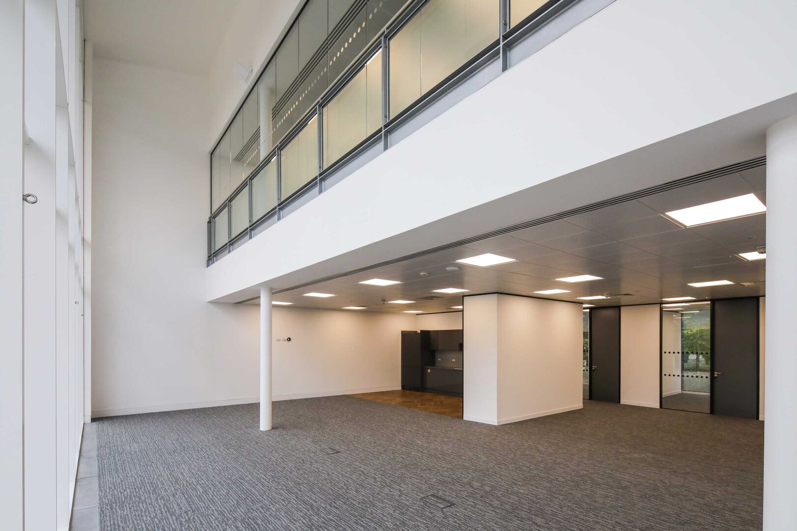 Modern, empty office space with carpeted floors, large windows, and a mezzanine level above.