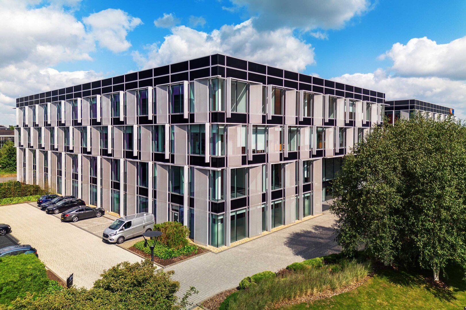 Modern glass office building with parked cars in front, surrounded by greenery under a partly cloudy sky.