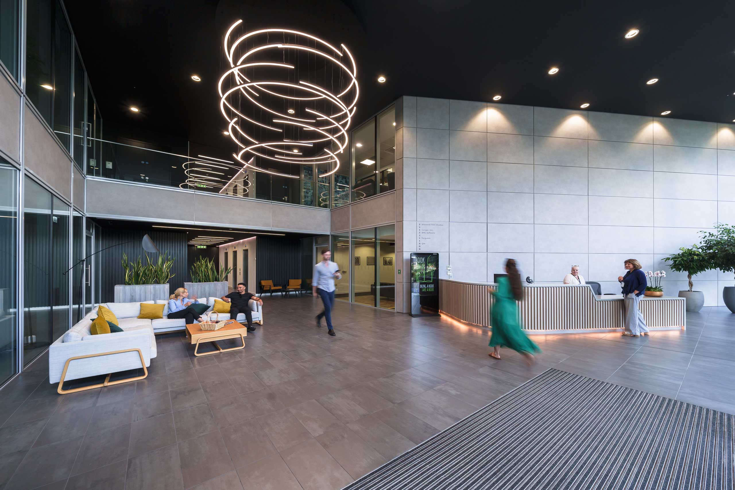 Modern office lobby with seating area, reception desk, and people walking or sitting in the space.