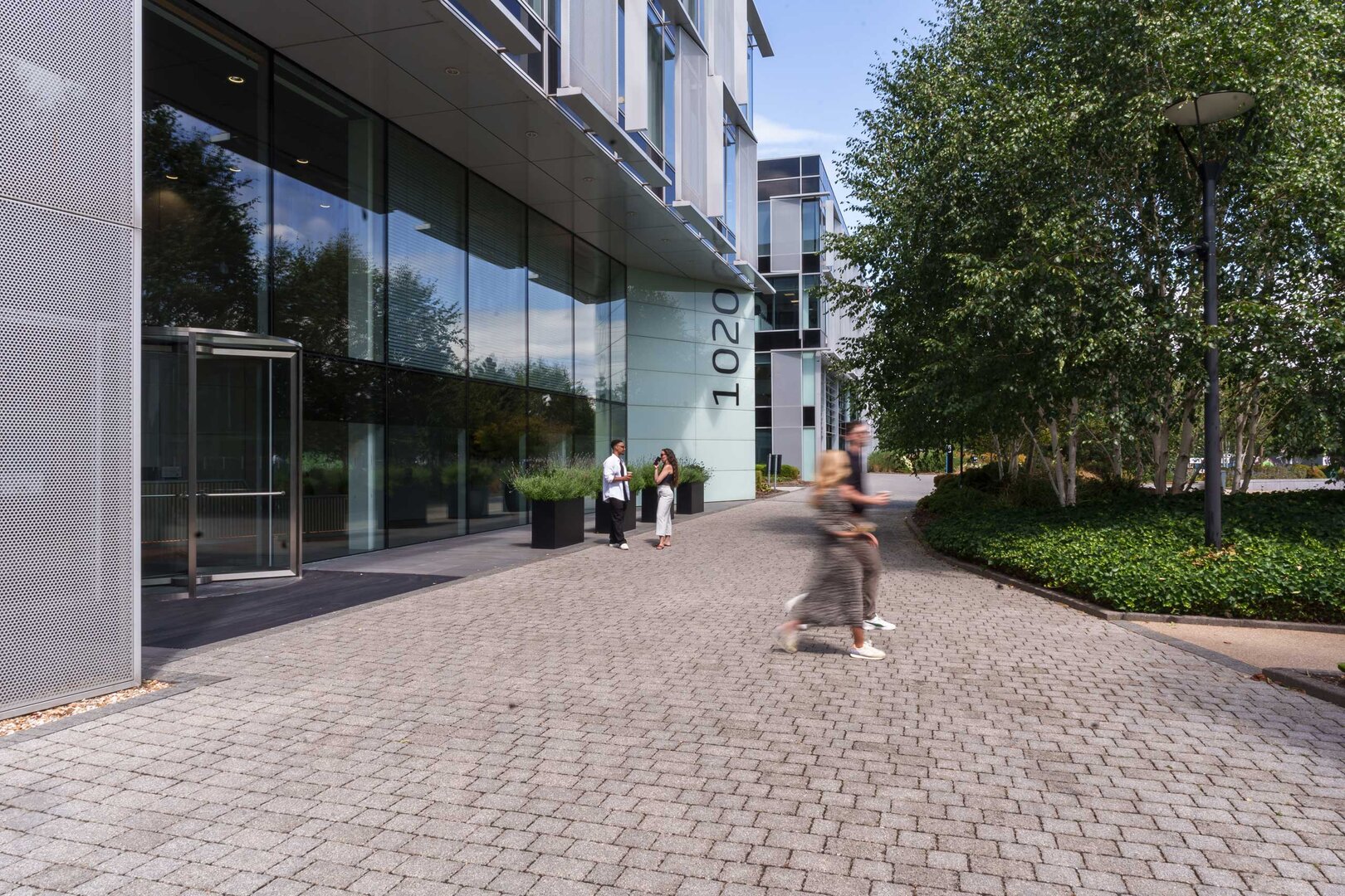 People walk outside a modern glass office building with the number 1020 on the entrance wall.