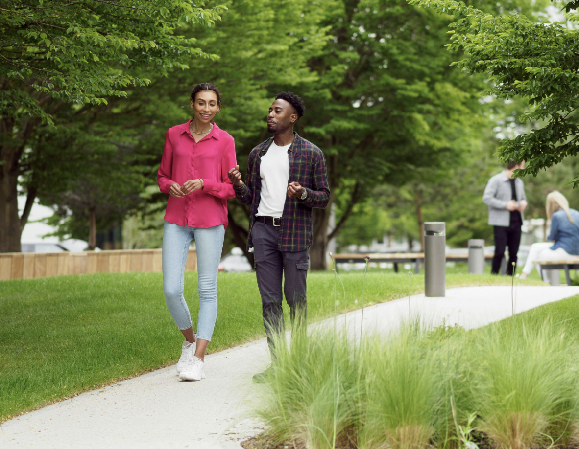 Two people walking and conversing on a paved path in a green park with benches and trees in the background.
