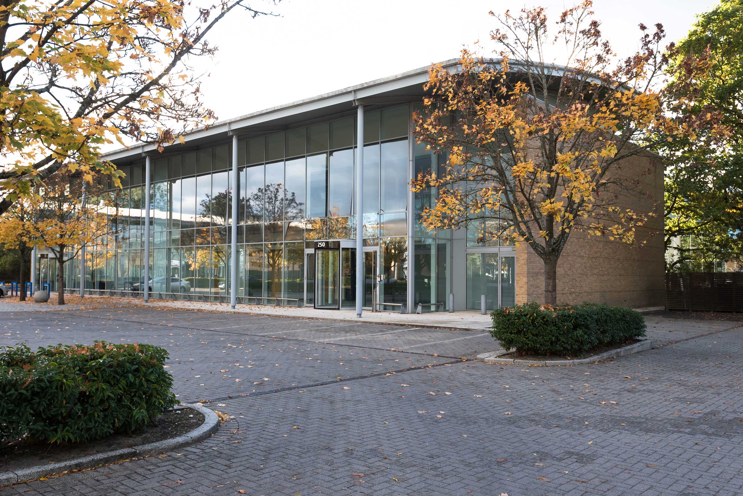 A modern building with large glass windows and autumn trees outside. The building is situated in a paved area with shrubs and no vehicles present.