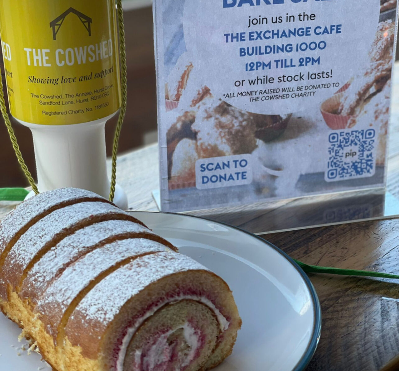 A rolled cake dusted with powdered sugar is on a plate in front of a bake sale sign and a donation box for The Cowshed charity.