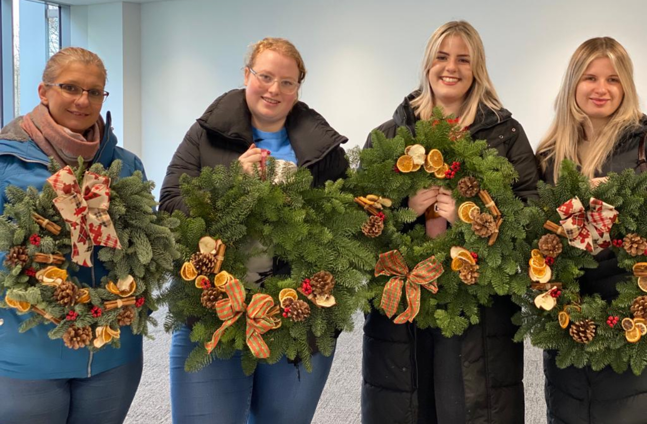Four people stand indoors holding decorative wreaths with pinecones and dried oranges.