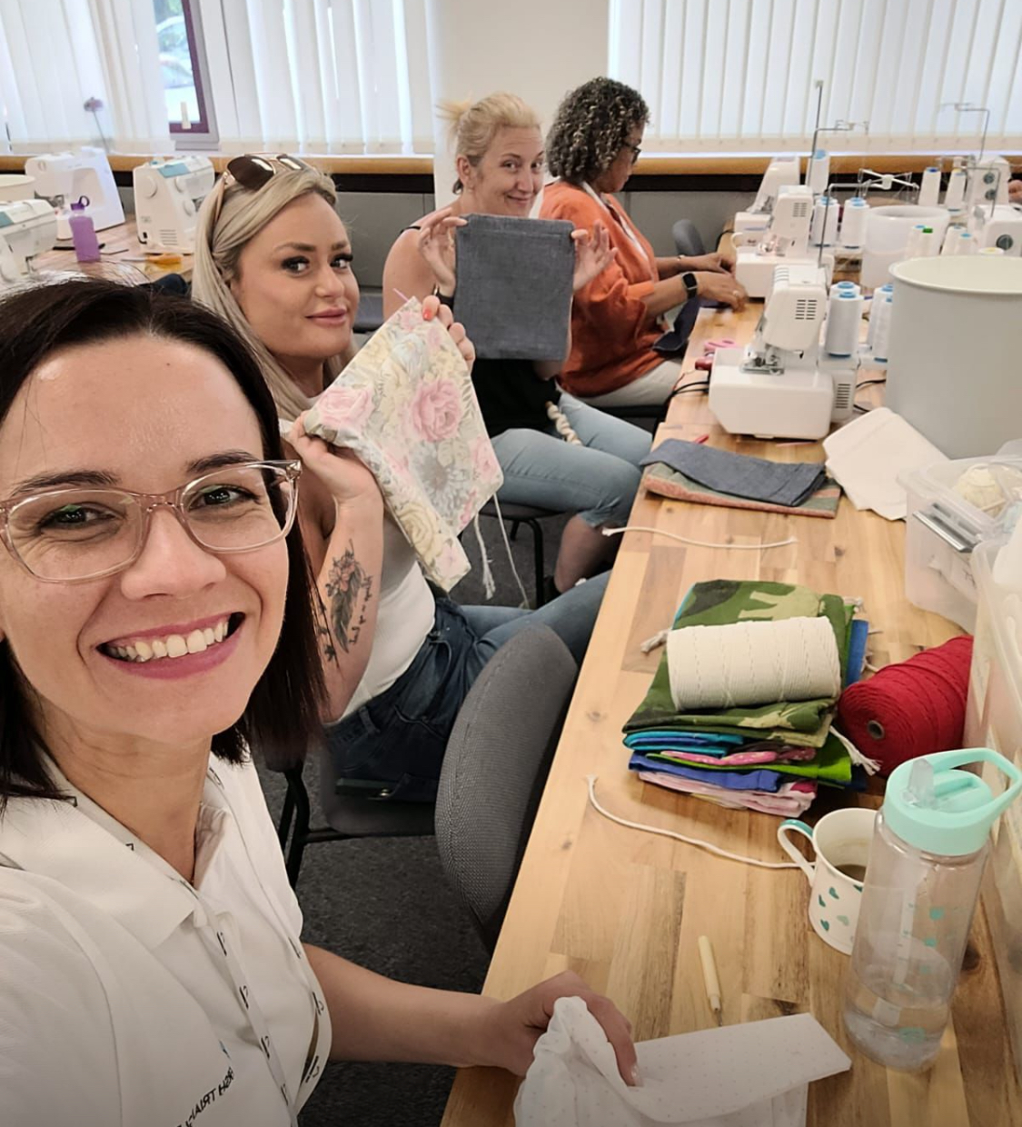 Four women sit at a long table with sewing machines, holding up fabric squares and smiling.