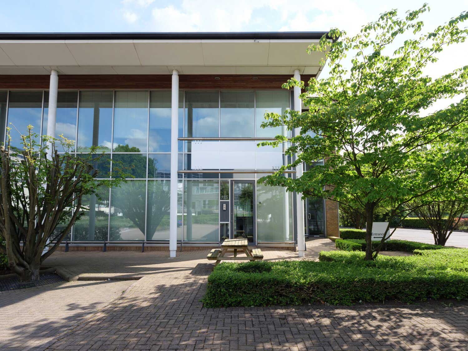 Modern glass office building with a picnic bench and green trees in the foreground.