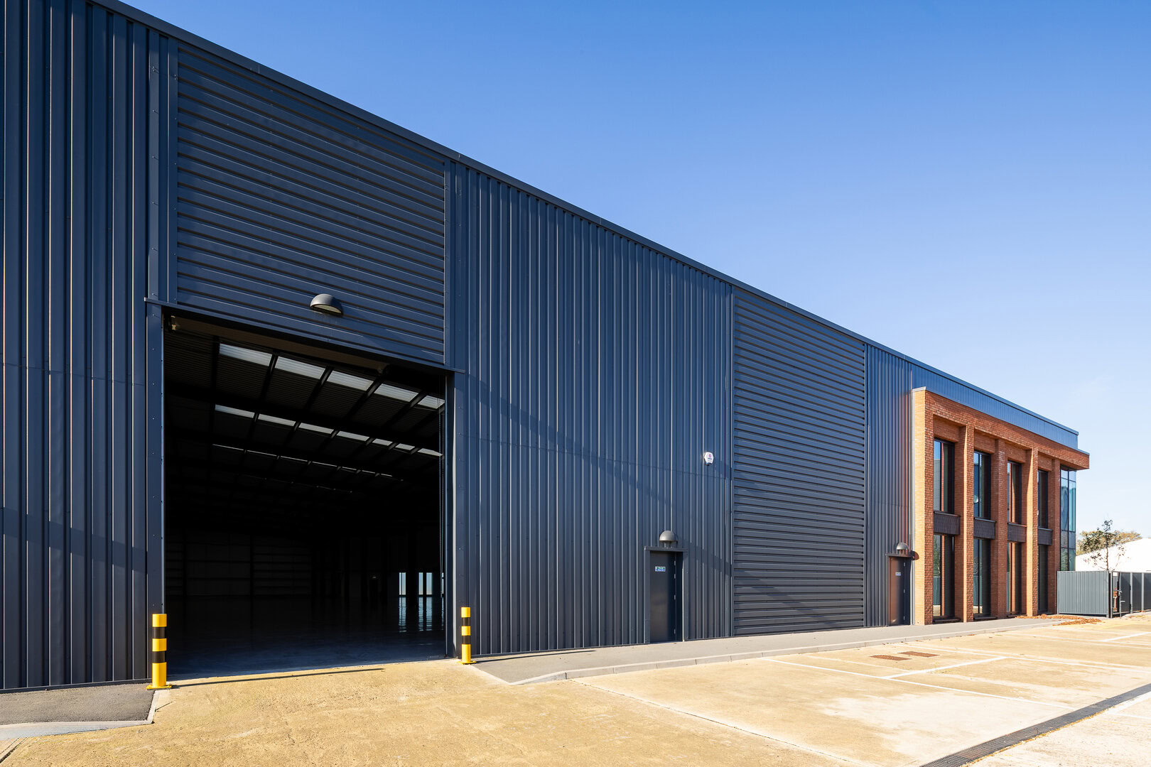 Large modern industrial warehouse with dark metal siding, open entrance, and adjacent parking area.