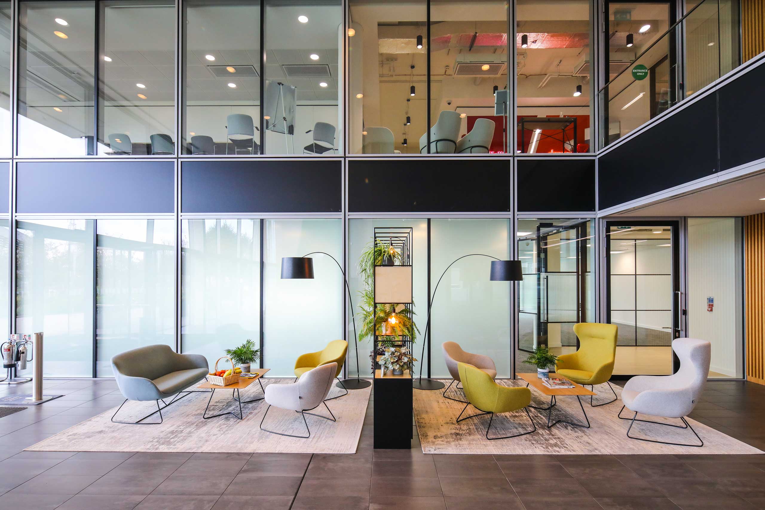 Modern office lobby with contemporary chairs, coffee tables, floor lamps, and glass walls; a mezzanine level with additional seating is visible above.