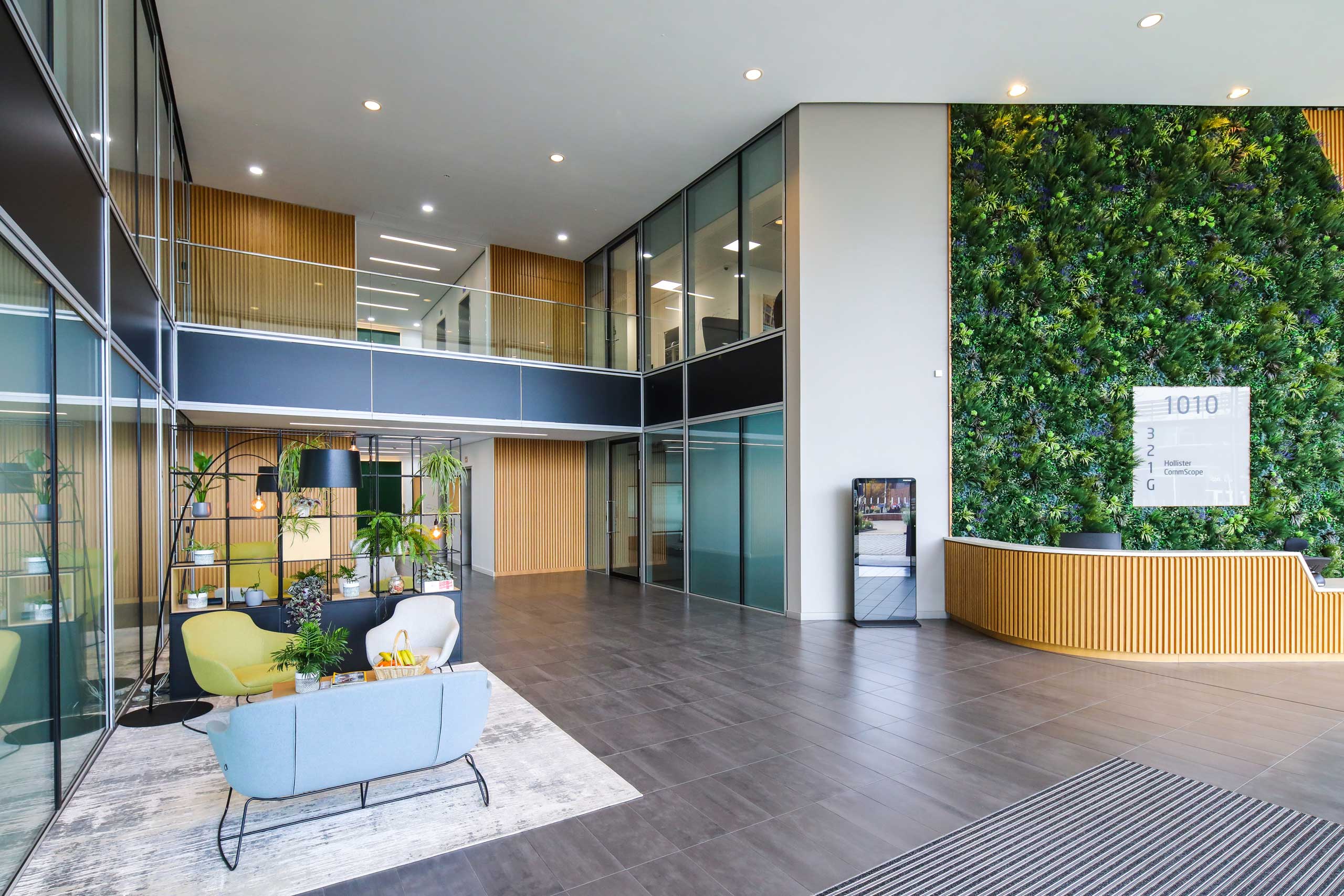 Modern office lobby with glass walls, seating area, a vertical green plant wall, wooden reception desk, and a directory sign displaying 1010.