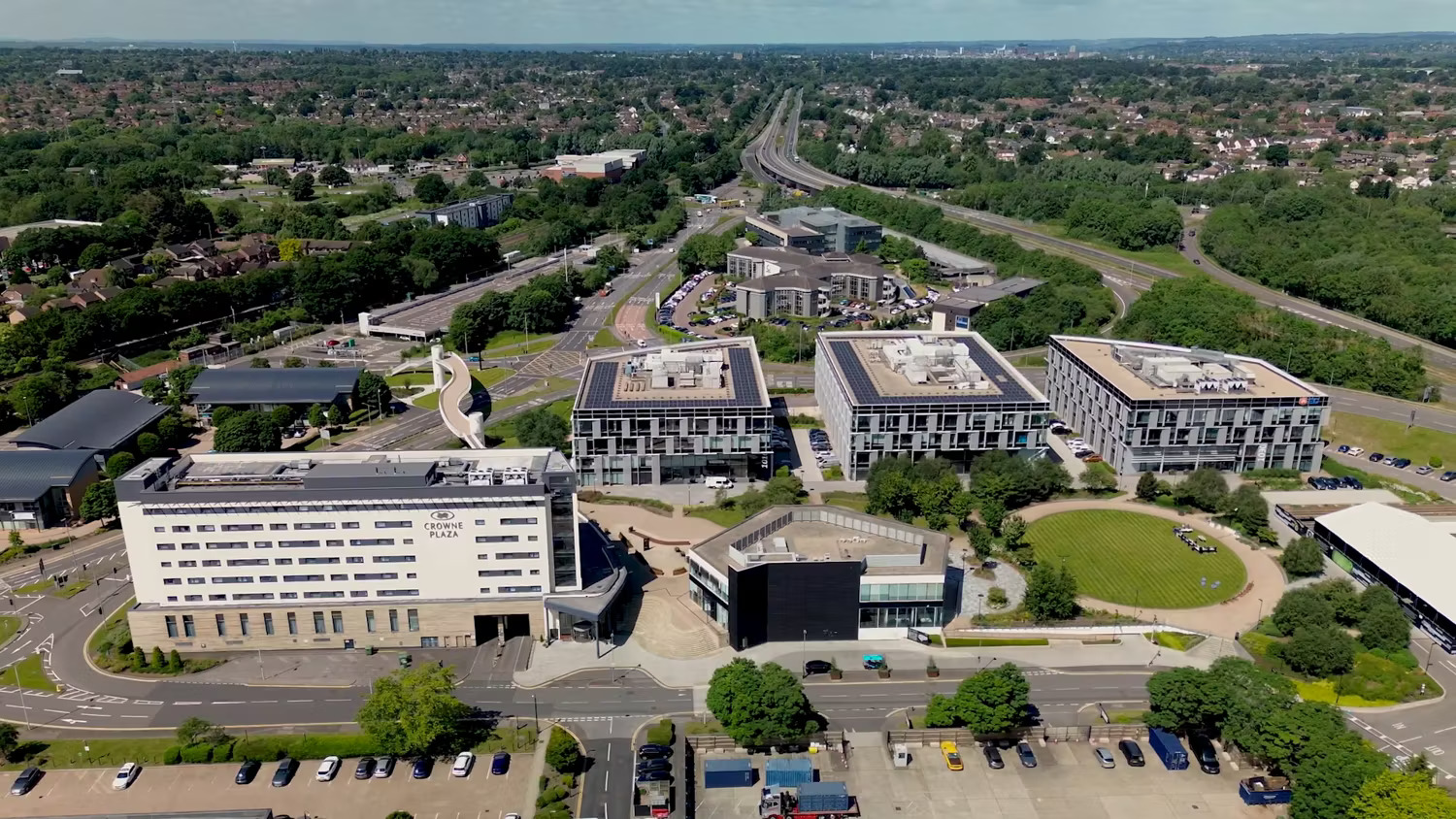 An aerial view of a modern office or academic complex with several rectangular buildings, a main road, green lawns, and car parks, surrounded by trees and a suburban area.