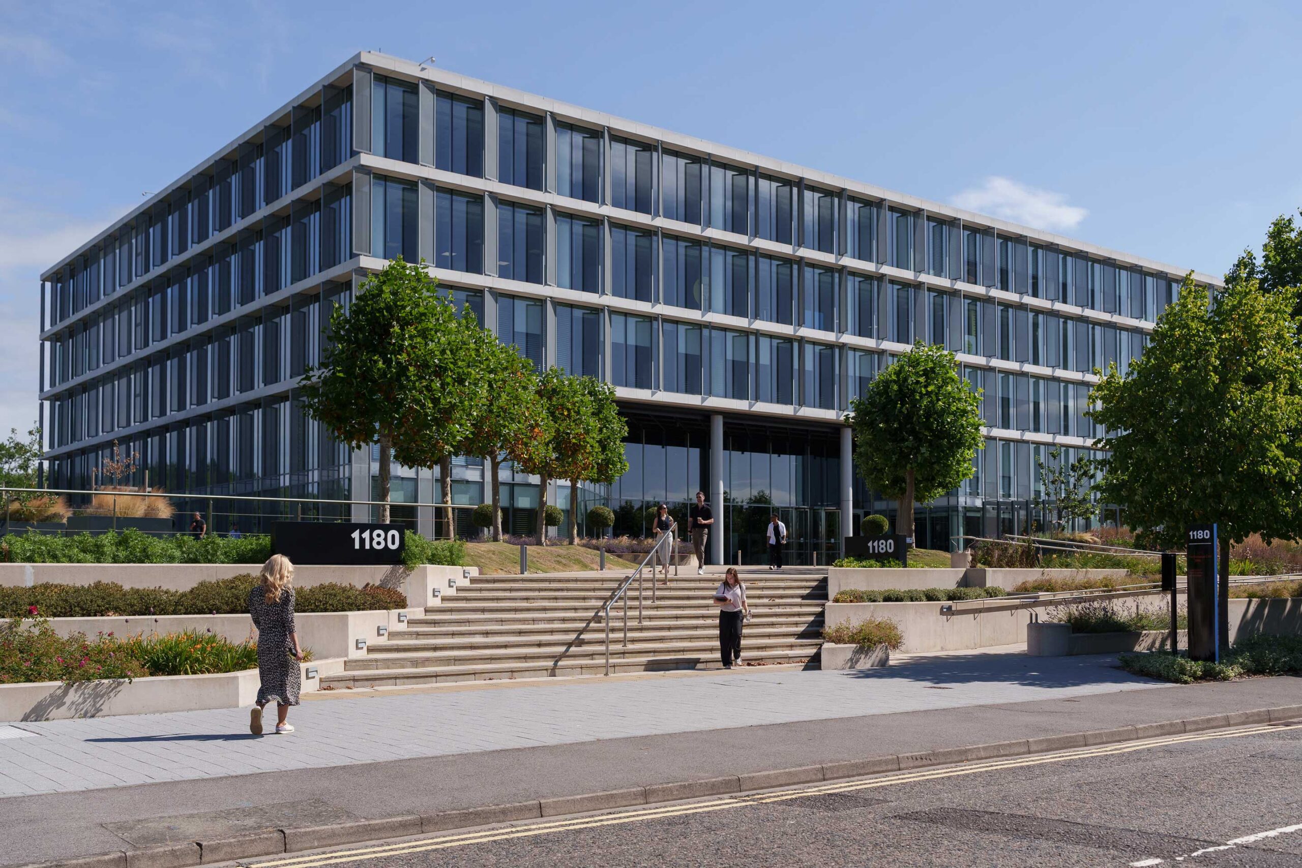 Modern glass office building with four storeys, small trees, and people walking near the entrance. The building number 1180 is visible on signs by the stairs.