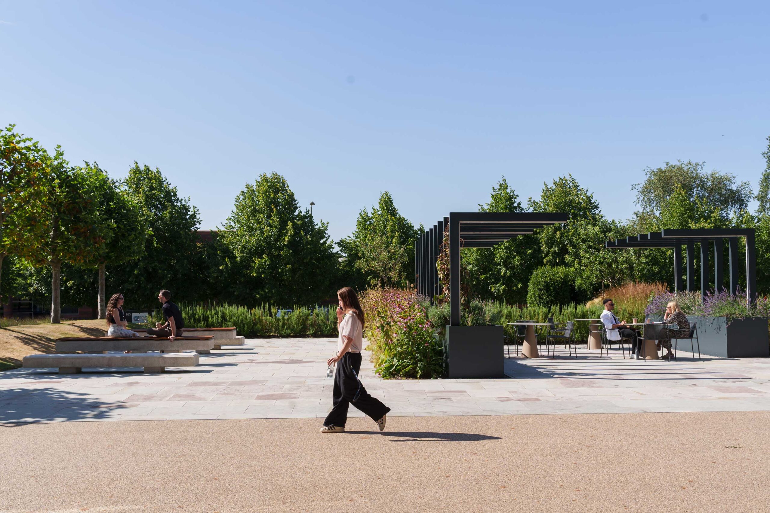 A person walks past a landscaped park area with trees, benches, and people sitting at outdoor tables on a sunny day.
