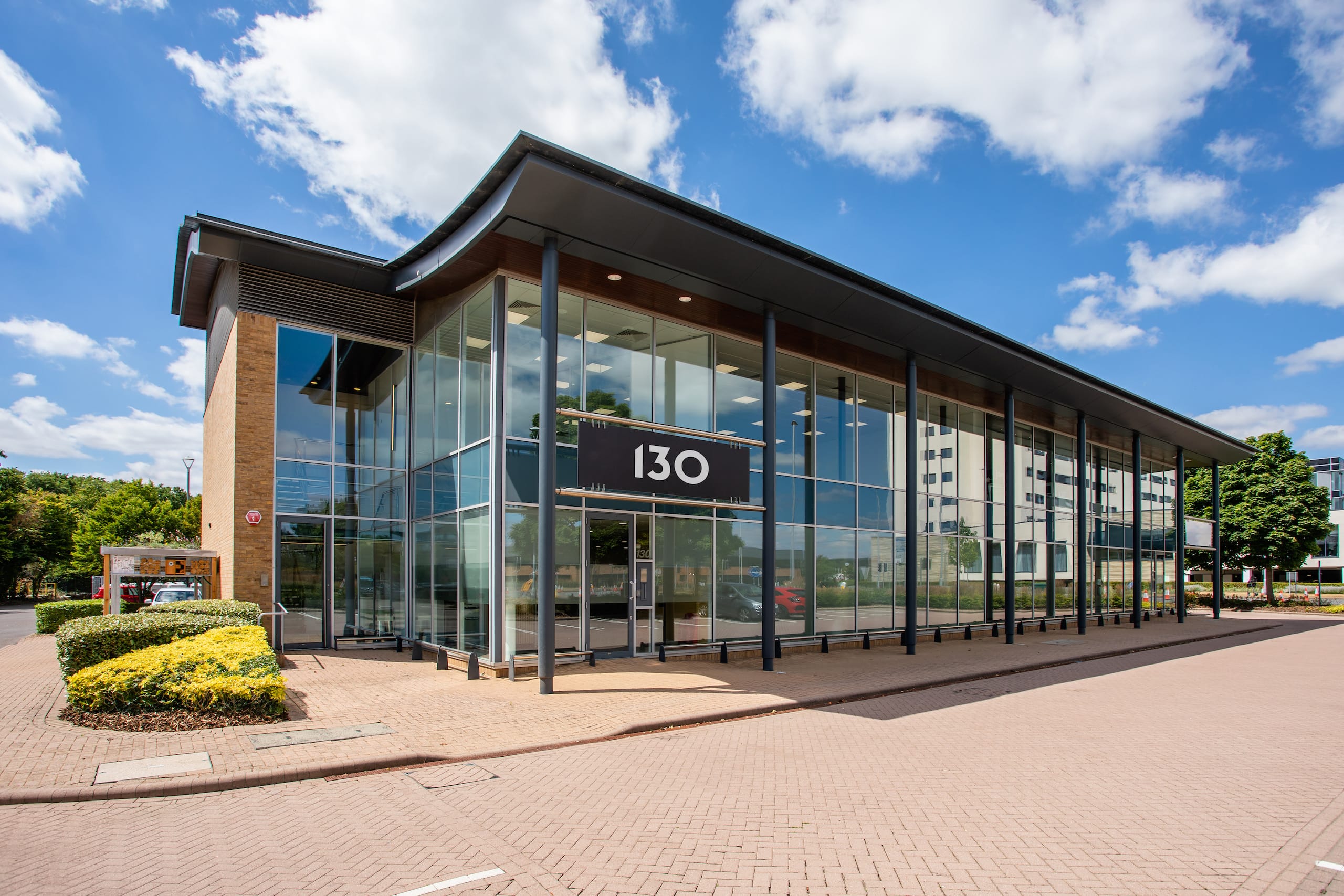 Modern two-storey glass building with a sign displaying “130” on the facade, set against a blue sky with scattered clouds. Paved area and landscaped bushes in front.