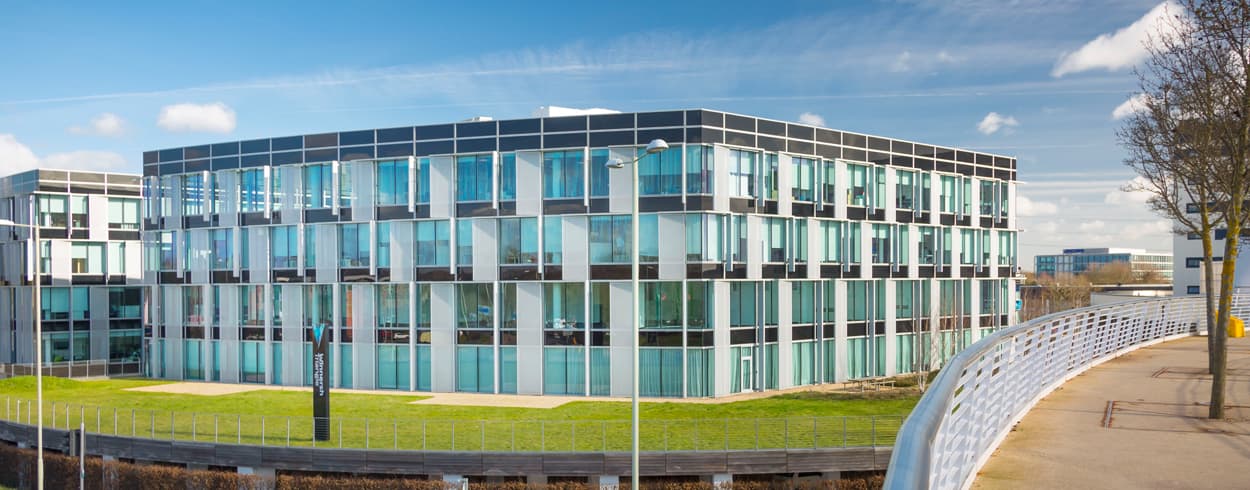 Modern glass office building with multiple storeys, large windows, and a geometric façade, situated next to a curved white walkway on a sunny day.