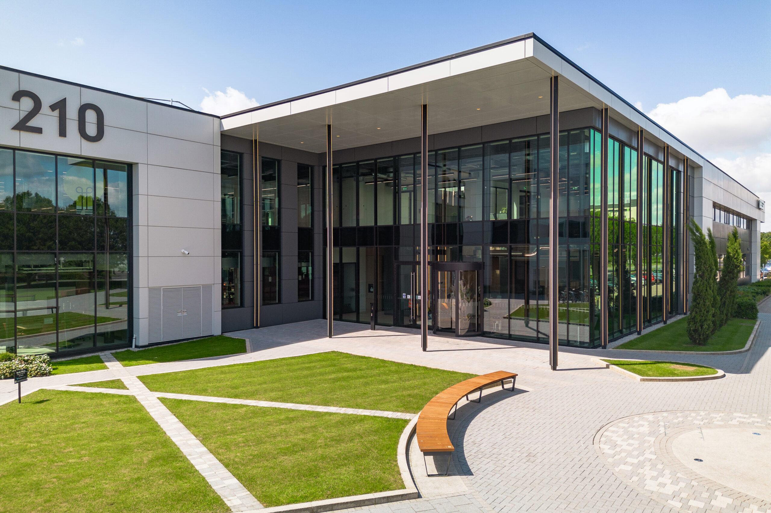 Modern office building with large glass windows, a landscaped lawn, pathway, curved bench, and the number 210 on the exterior wall.