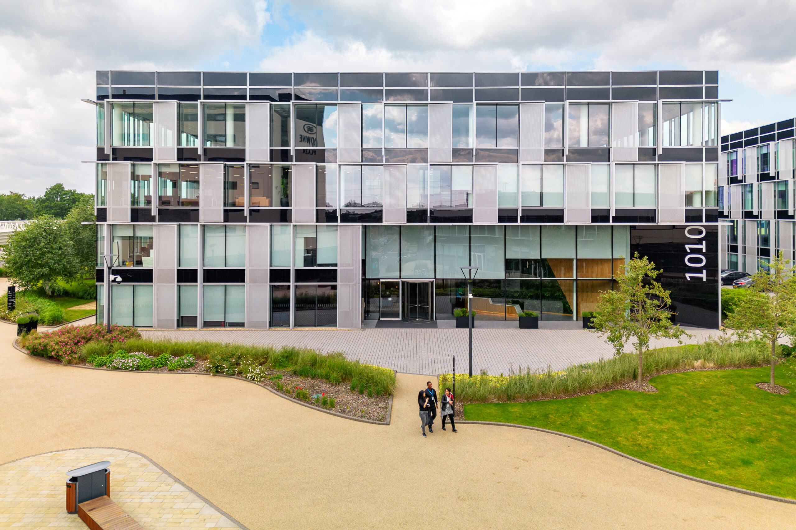 Modern glass office building with the number 1010 on the side, surrounded by landscaped paths and greenery. Three people walk on a path in front of the building.