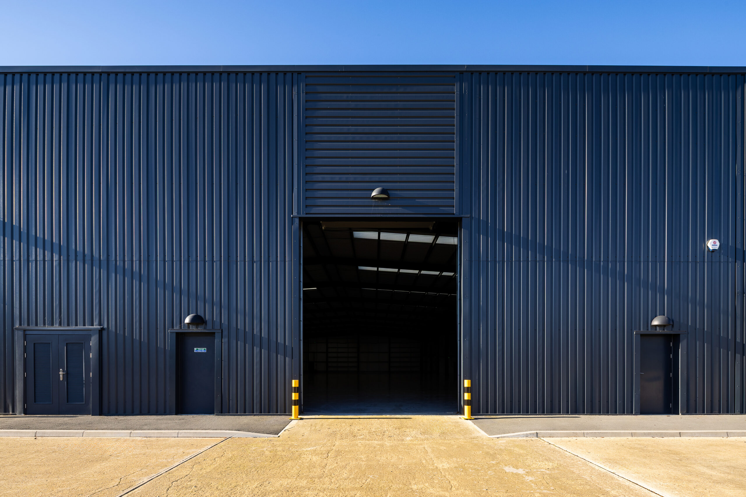 Exterior of a large industrial warehouse with dark metal cladding, an open central entrance, and two closed doors on either side, under a clear blue sky.