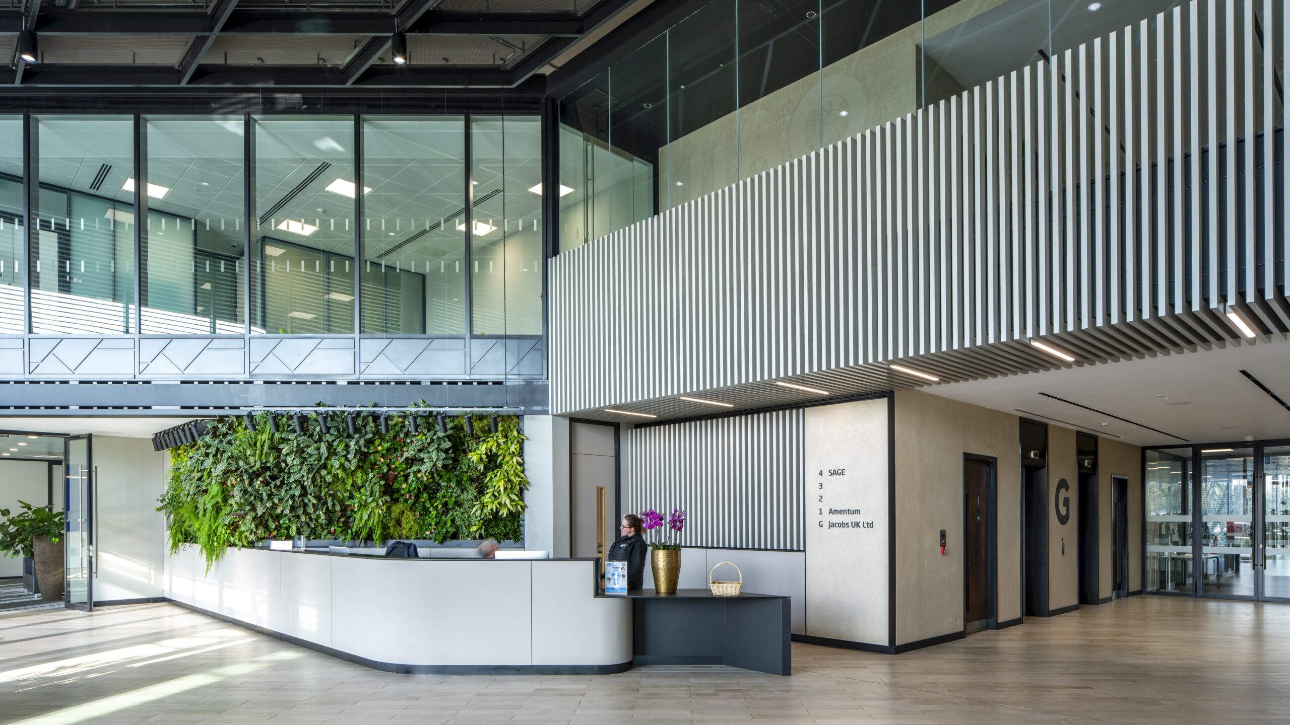 Modern office lobby with a vertical garden behind the reception desk, directional signage on the wall, lifts to the right, and glass-walled offices above.