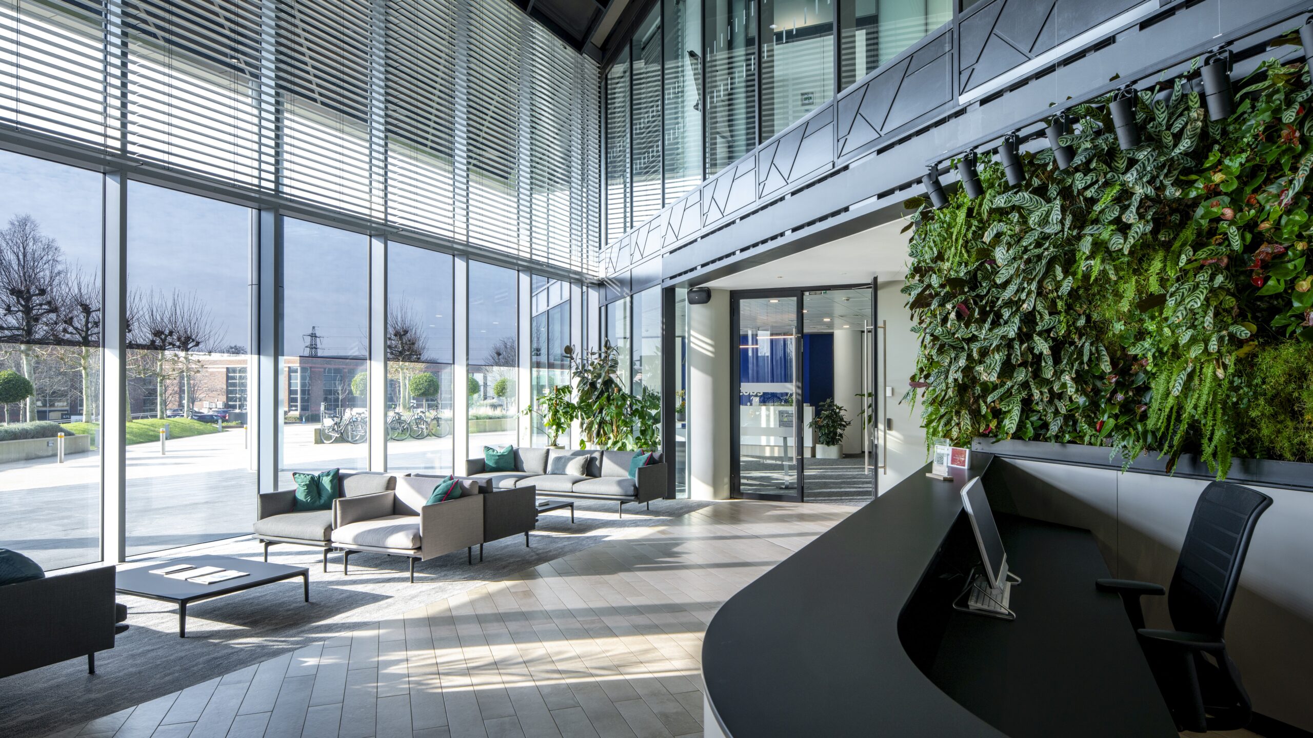 Modern office lobby with large glass windows, indoor plants, living wall, reception desk, and seating area with sofas and chairs.