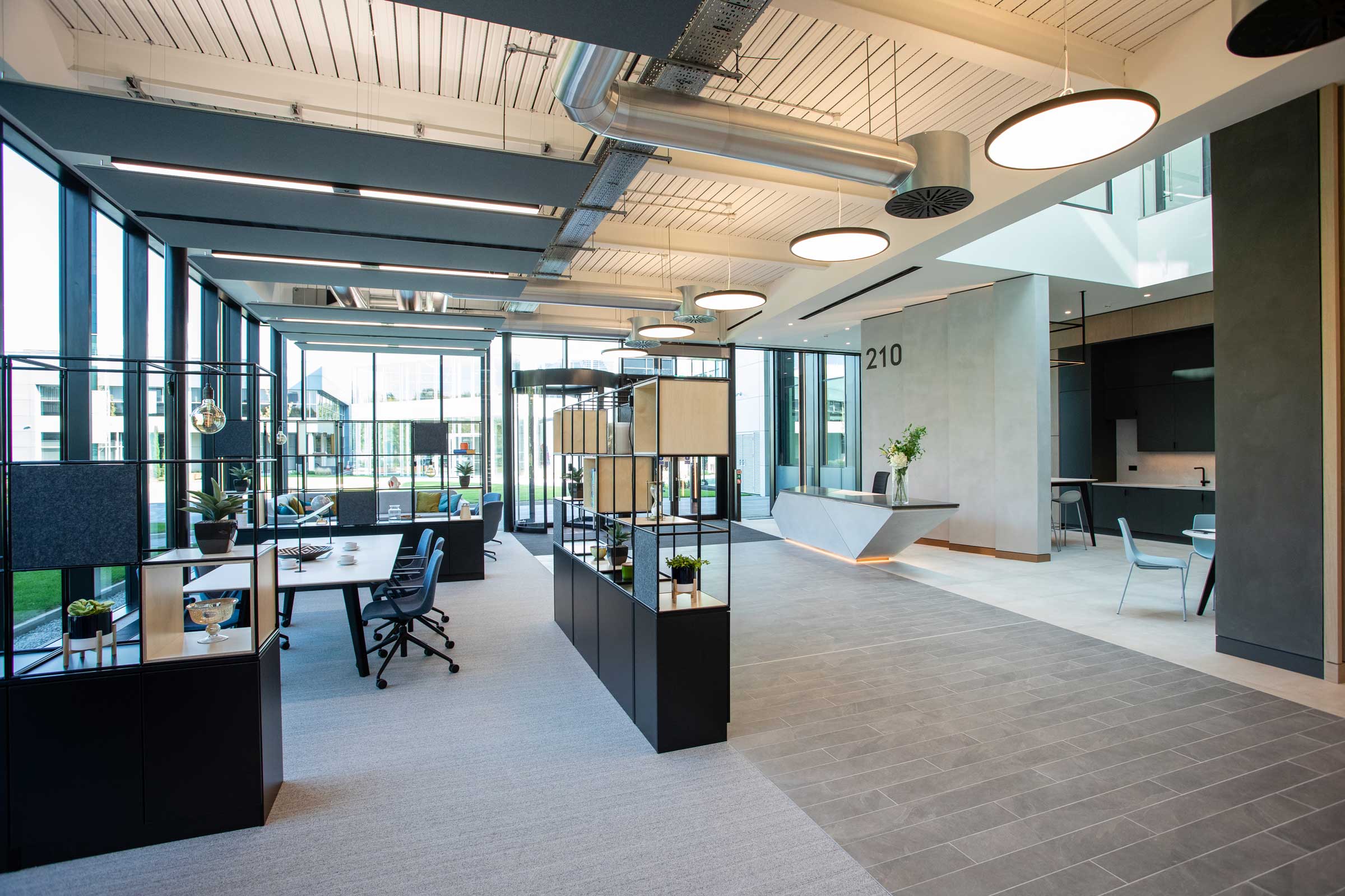 Modern office space with large windows, open workstations, plants, and a reception area; bright lighting and contemporary furnishings throughout.