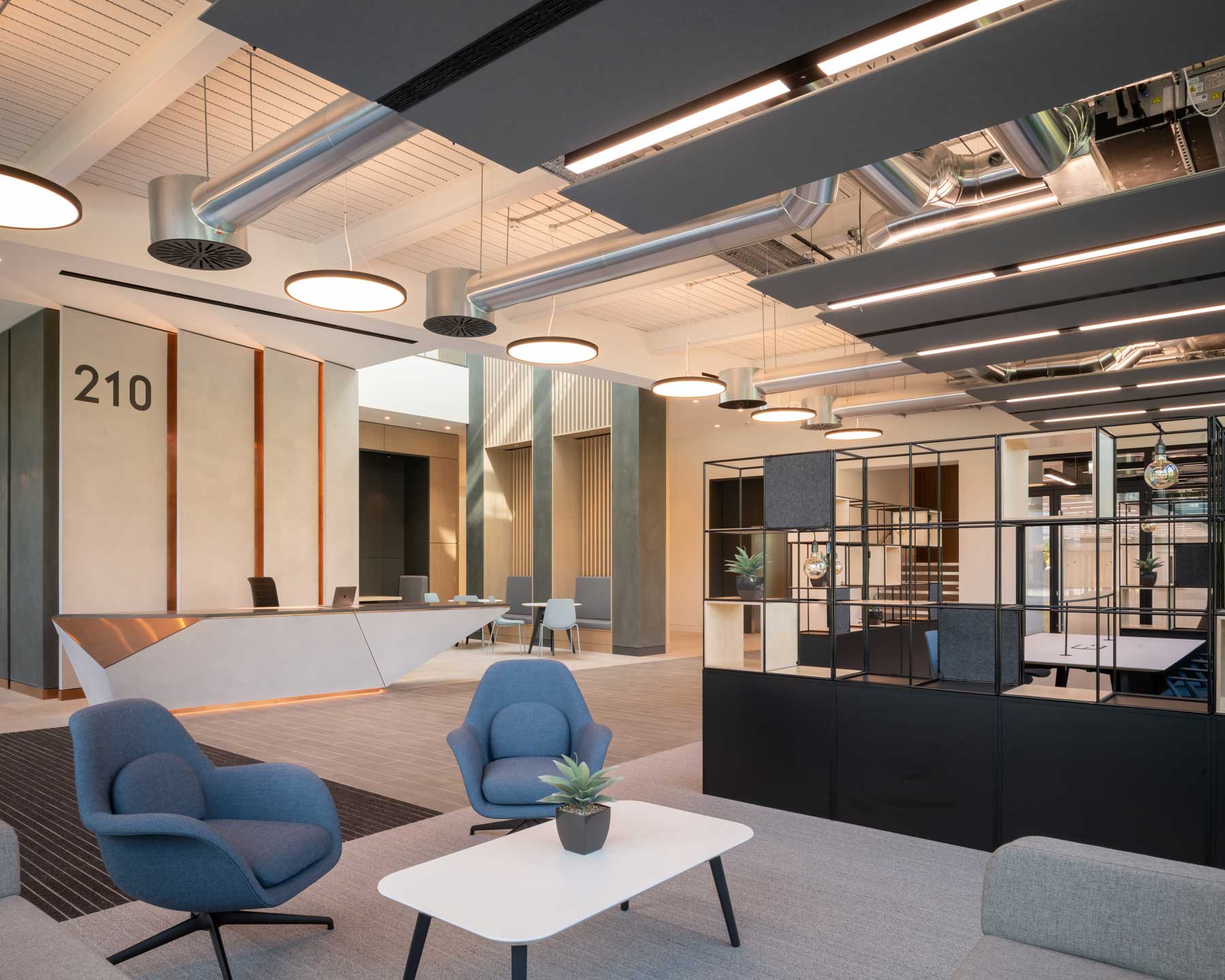 Modern office lobby with a geometric reception desk, numbered wall, lounge chairs, tables, and exposed ceiling ducts.
