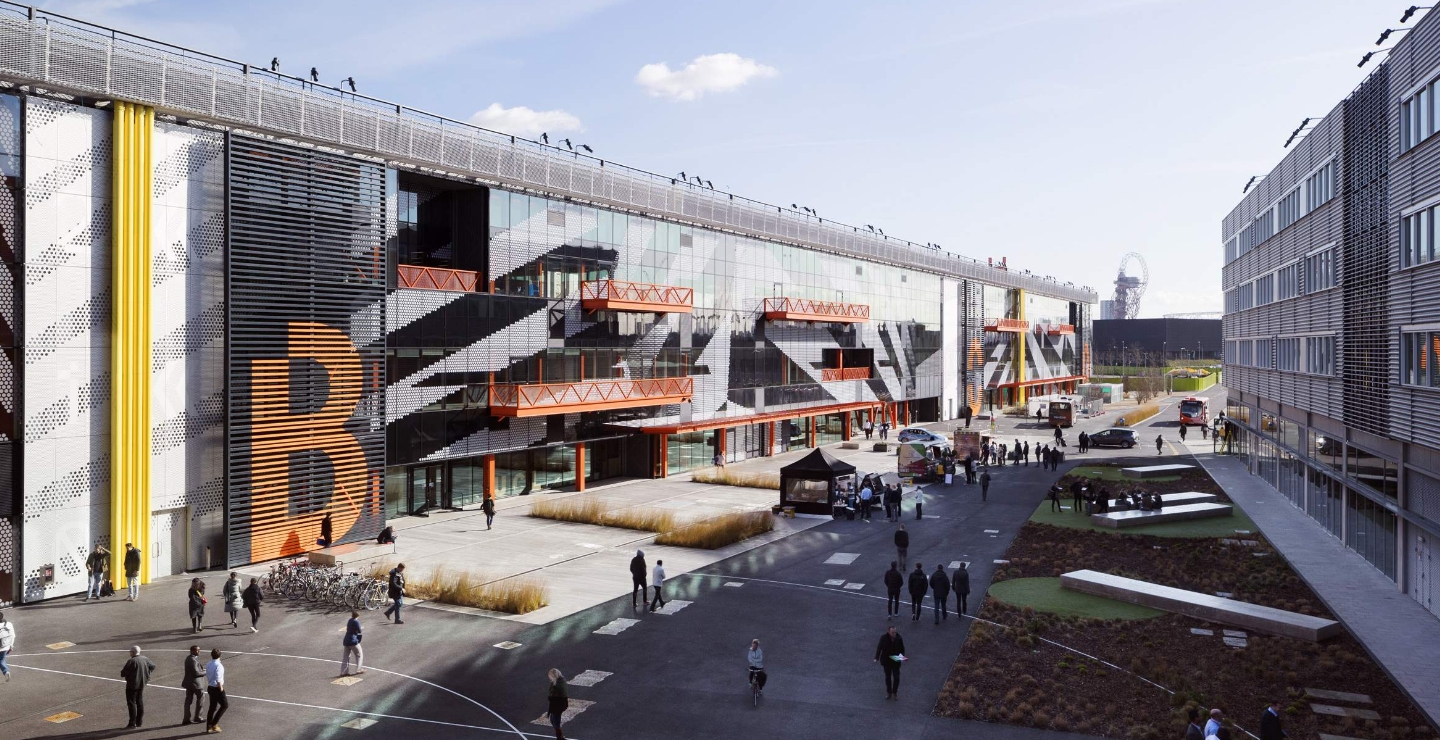 Modern multi-storey building with large glass windows and orange accents, people walking outside, landscaped courtyard, and clear sky overhead.