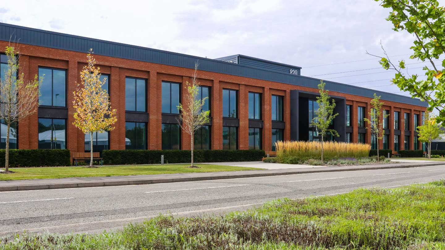 A modern two-storey brick office building with large windows, young trees, and landscaped greenery along an empty street under a cloudy sky.