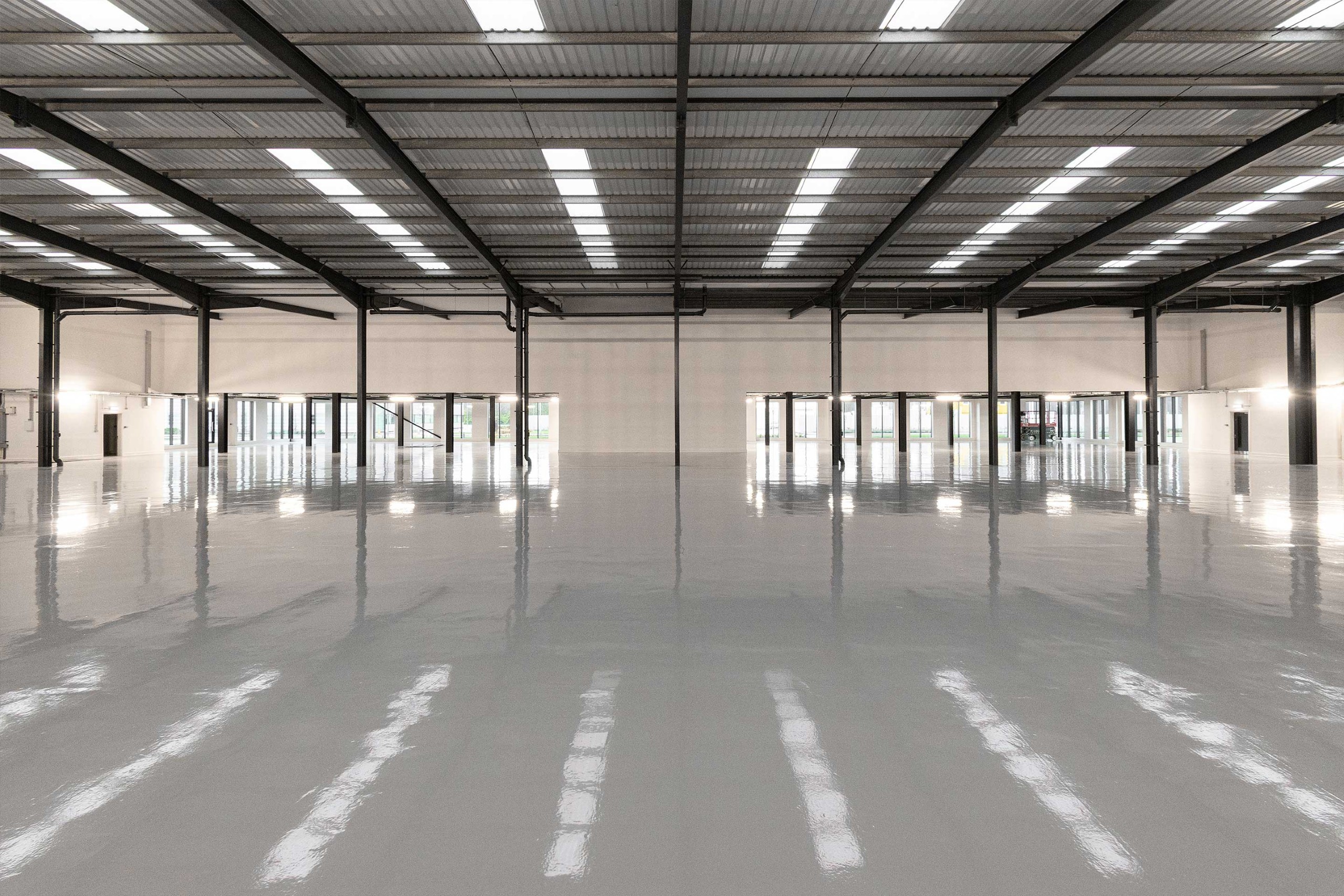 Large, empty warehouse interior with polished concrete floor, exposed steel beams, and rows of ceiling lights. Windows and doors are visible along the far wall.