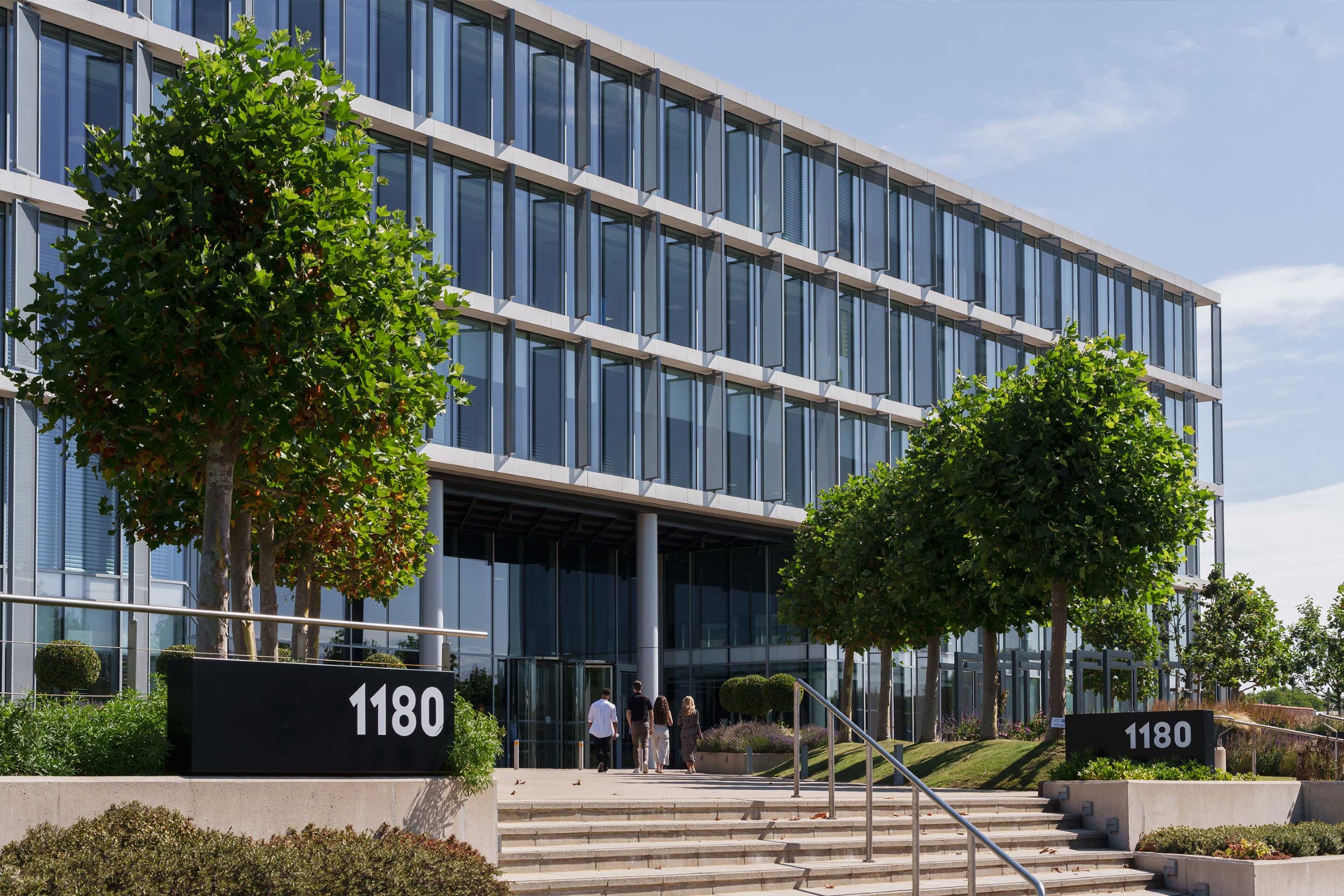 Modern glass office building with the number 1180 displayed on signs near the entrance; several people are walking towards the main doors.