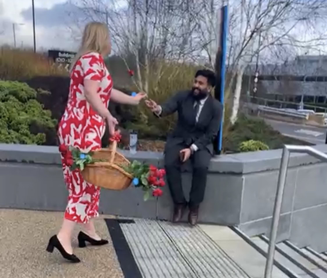 Woman in a red dress hands a rose to a seated man in a suit outdoors by some steps.