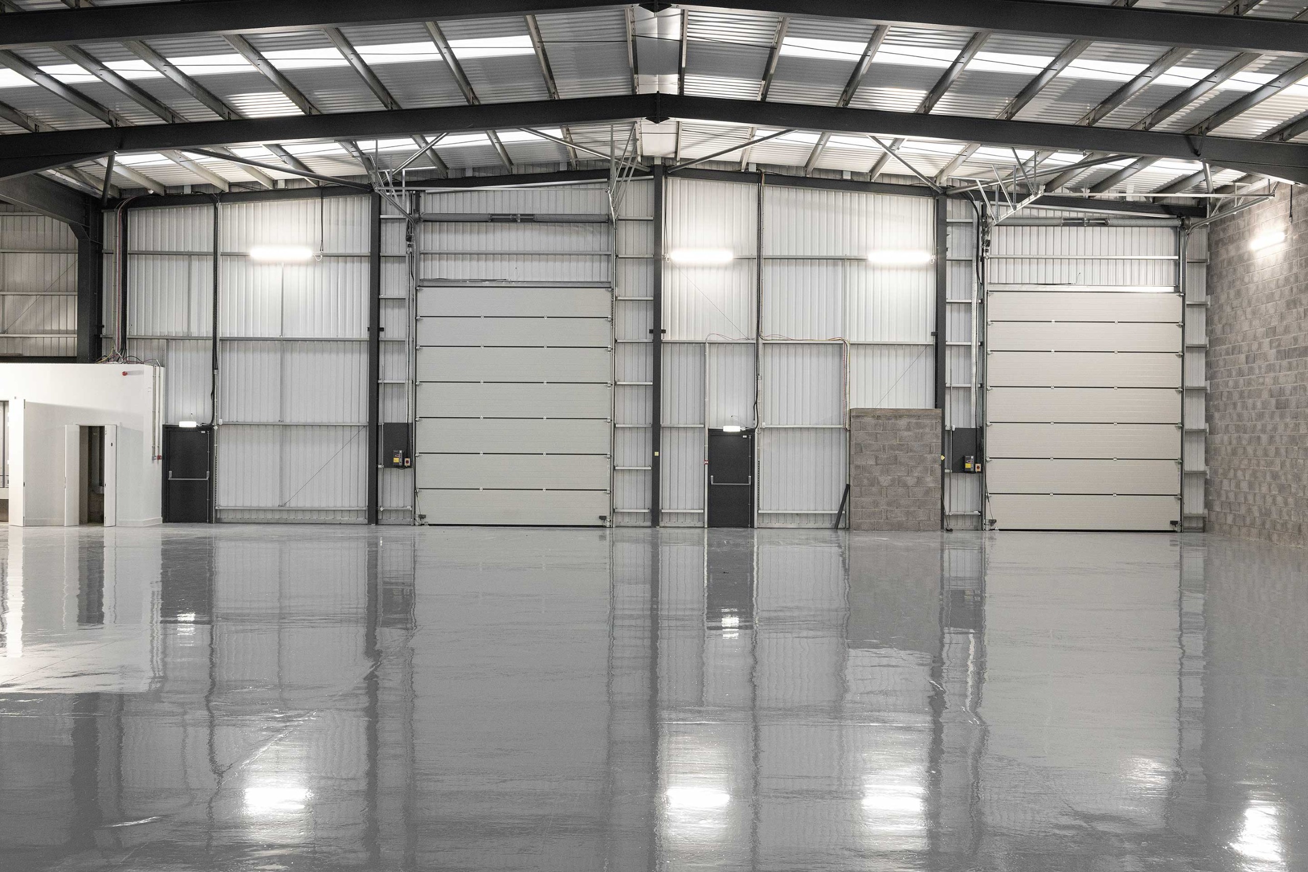 Interior of an empty industrial warehouse with high ceilings, metal walls, large roller doors, and a polished concrete floor.