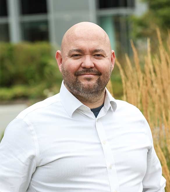 A bald man with a beard wearing a white shirt stands outdoors in front of greenery and a building, smiling at the camera.