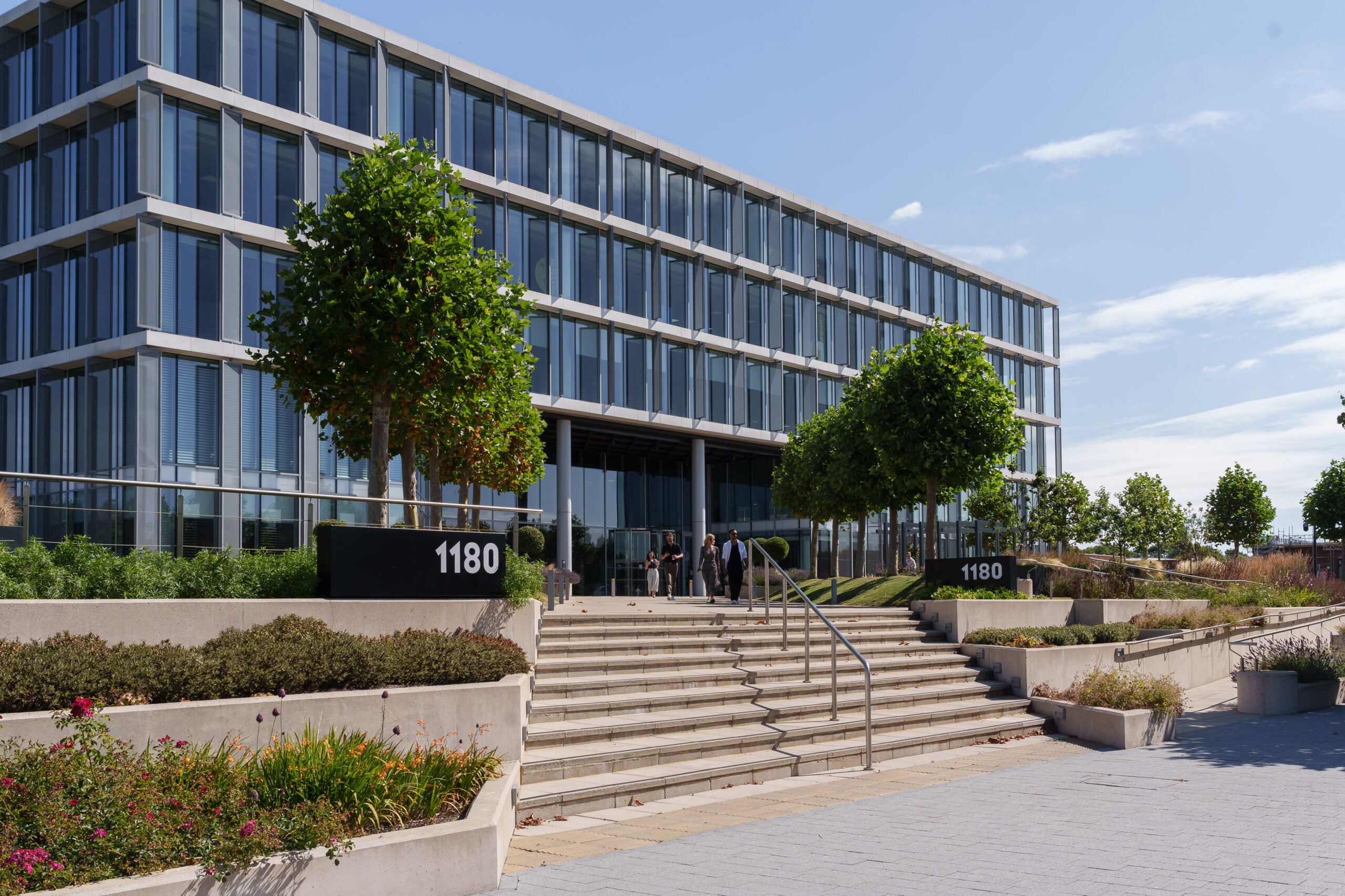 Modern glass office building with stairs, landscaping, and prominent 1180 address markers; several people stand near the entrance under a clear sky.