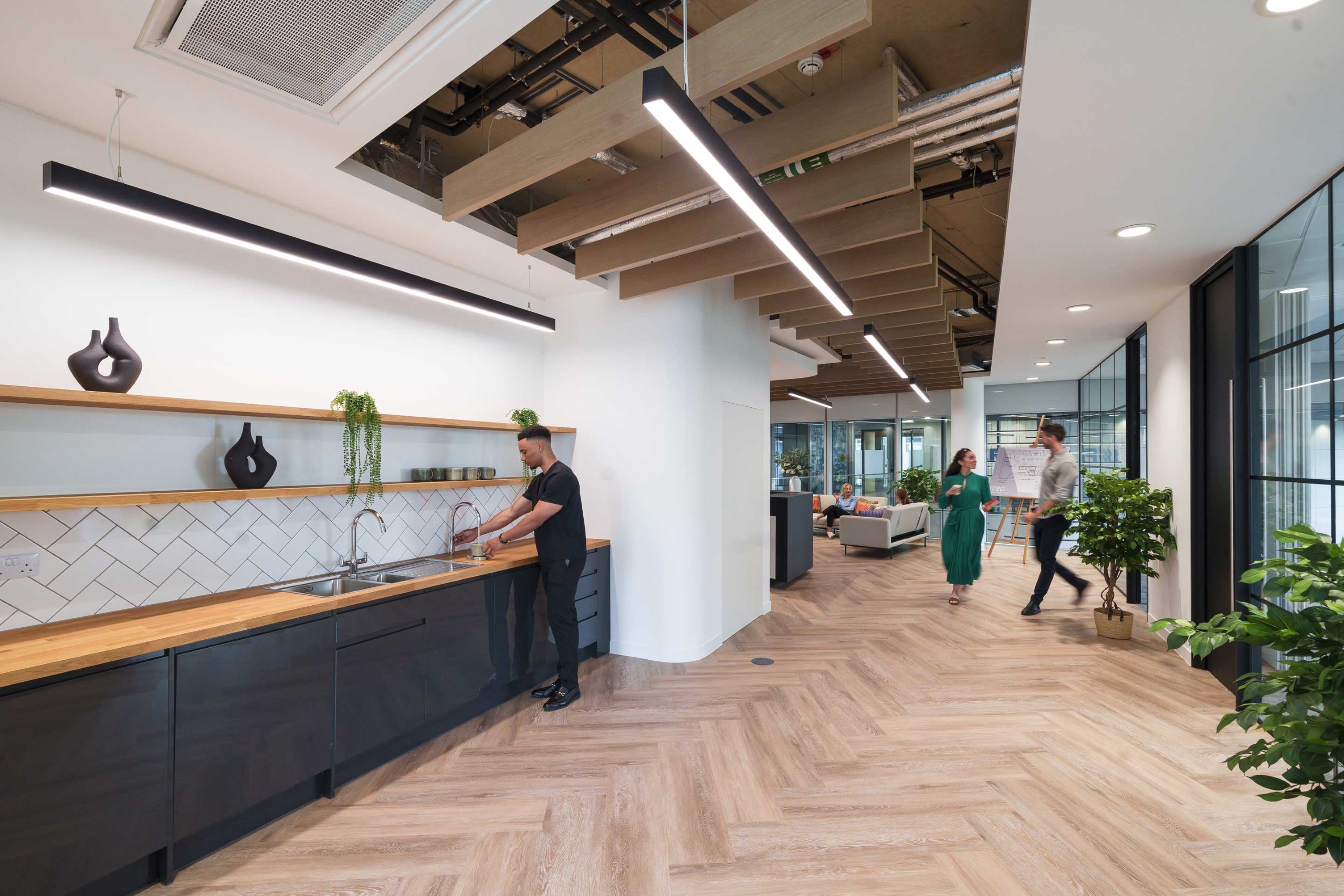 A man washes dishes at a kitchen sink in a modern office space, whilst two people walk and talk in the background.