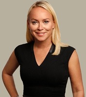 A woman with straight blonde hair, wearing a black sleeveless top, stands against a plain beige background, smiling at the camera.