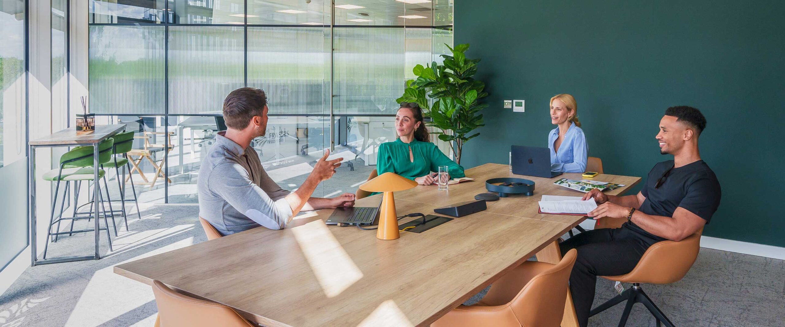 Four people sit around a conference table in a modern office, engaged in discussion, with laptops, notebooks, and water glasses on the table.