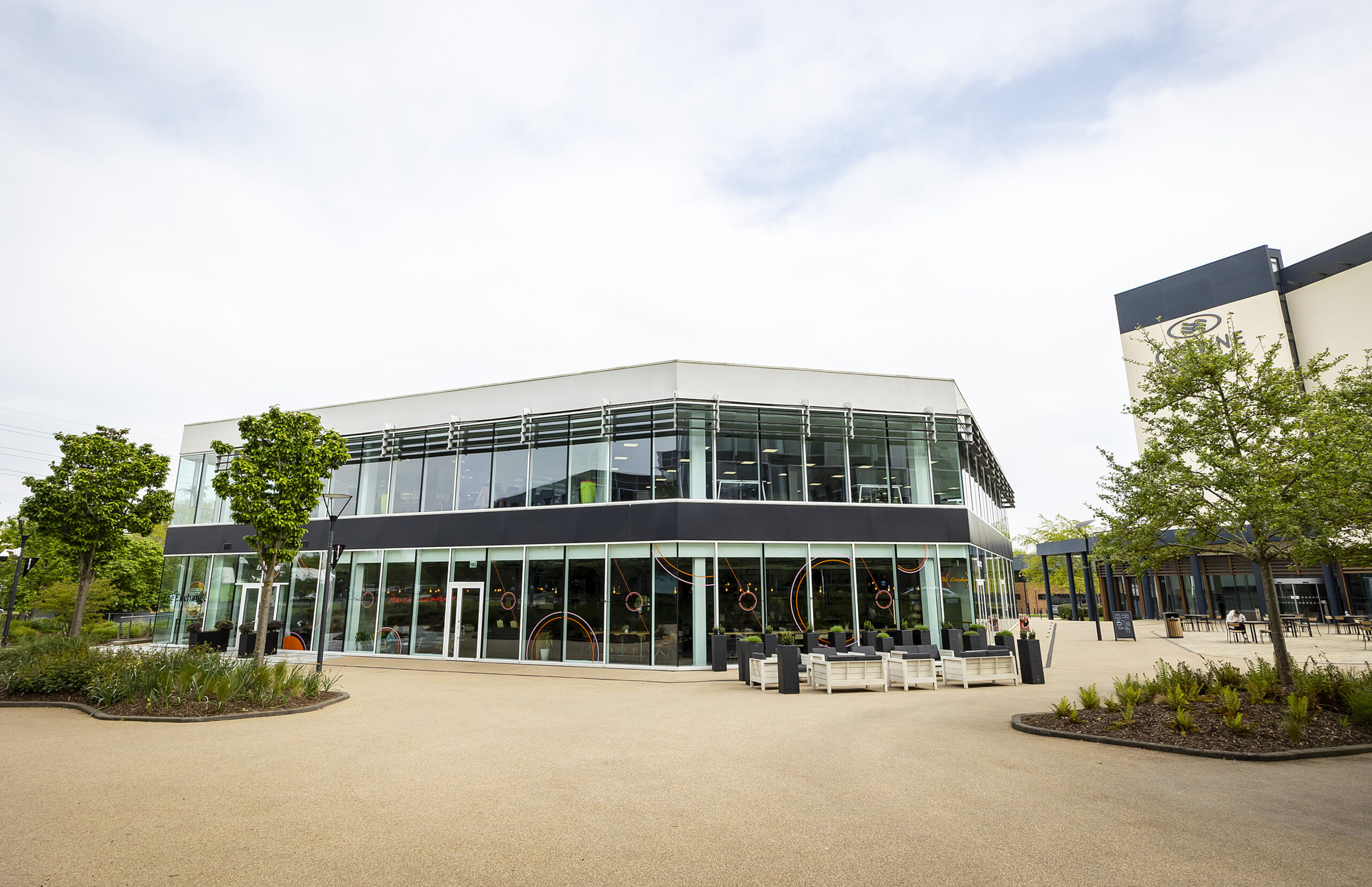 Modern two-storey glass building with outdoor seating and landscaping, photographed on a cloudy day.