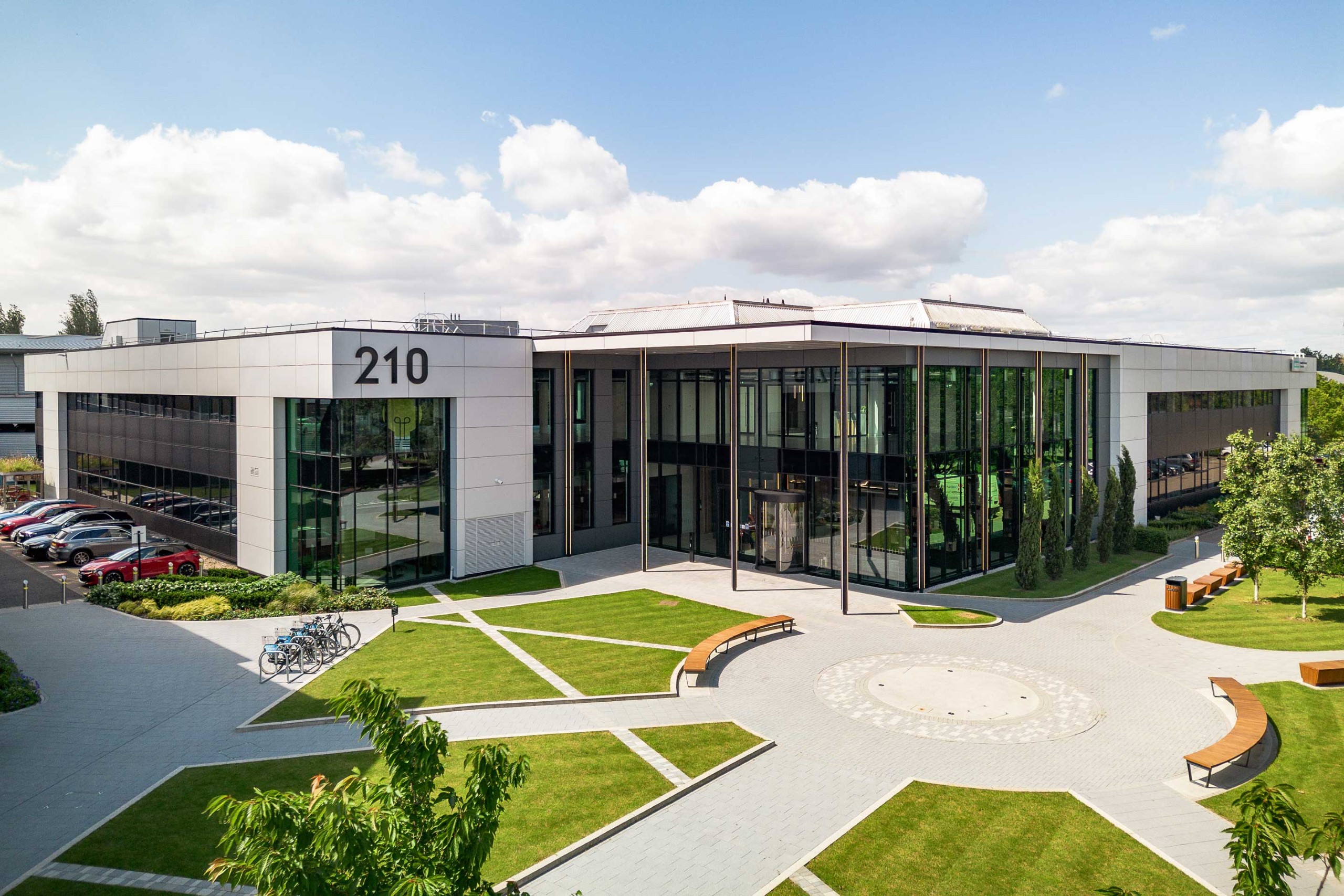 Modern office building with large glass windows, labelled 210, surrounded by green lawns, benches, trees, and parked cars under a partly cloudy sky.