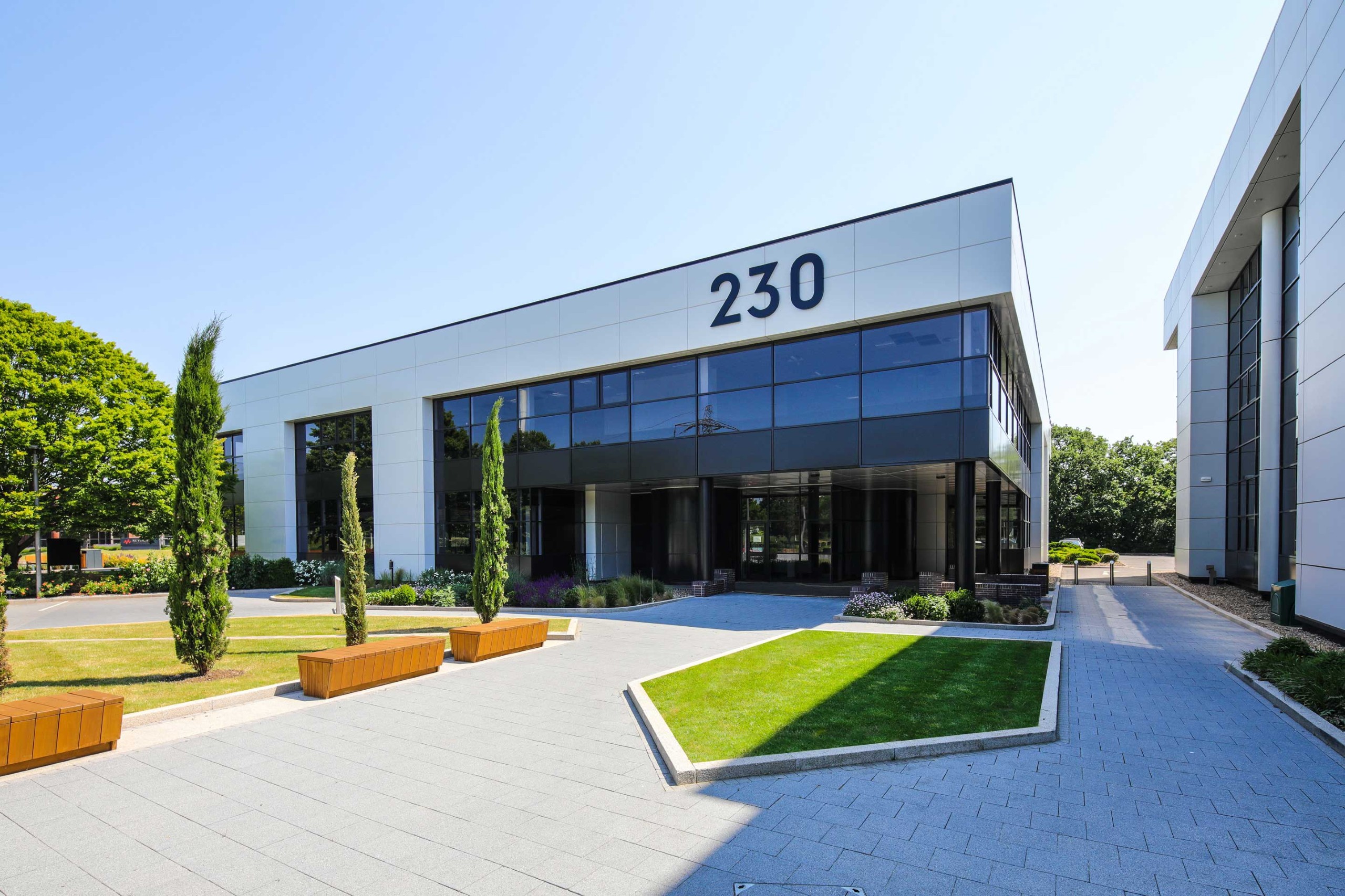 Modern office building with large windows and the number 230 displayed above the entrance, surrounded by trees and landscaping on a sunny day.