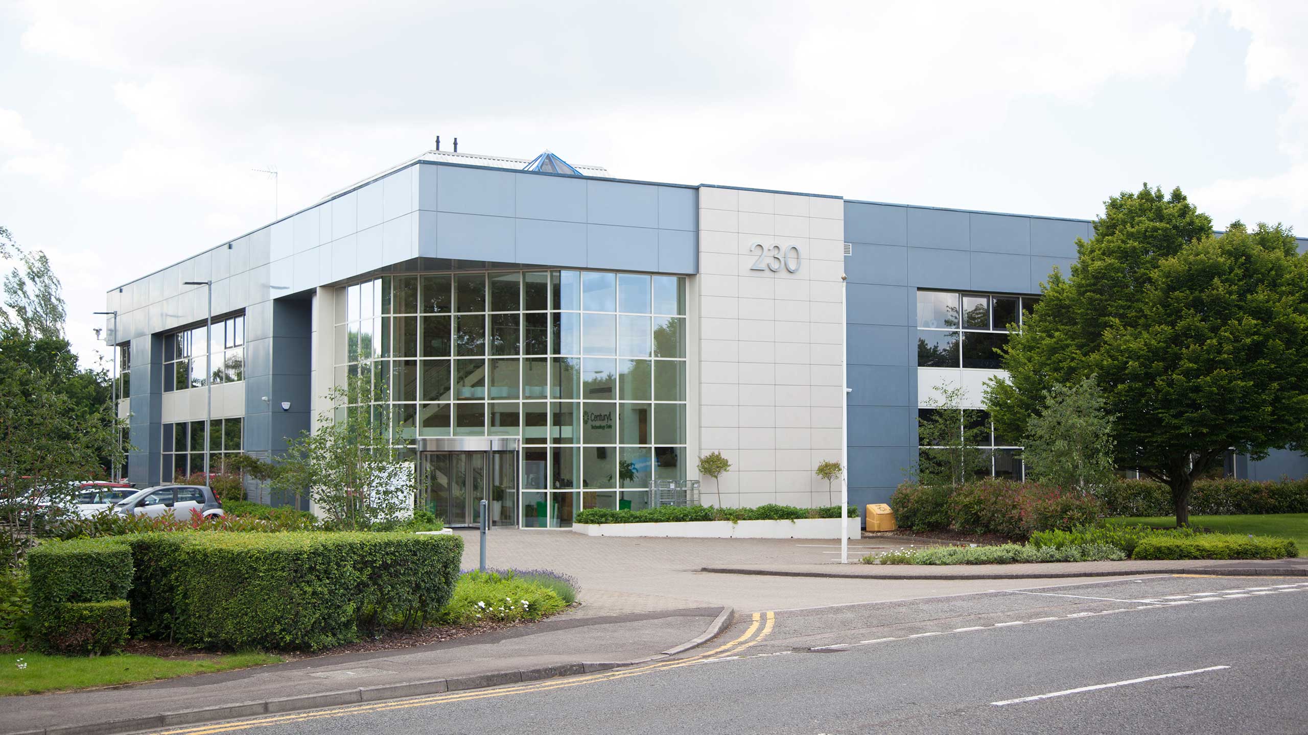 Modern two-storey office building with large windows and number 230 on the facade, surrounded by greenery and a car park, viewed from across the street.