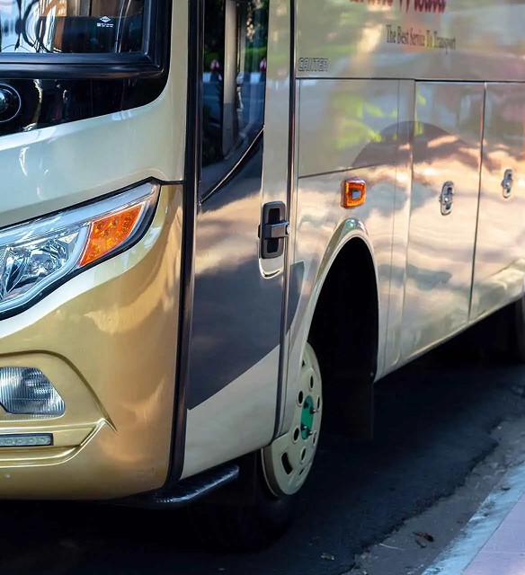 Close-up of the front and side of a parked beige coach with green wheel hub, showing part of the headlamp, door, and side panels.