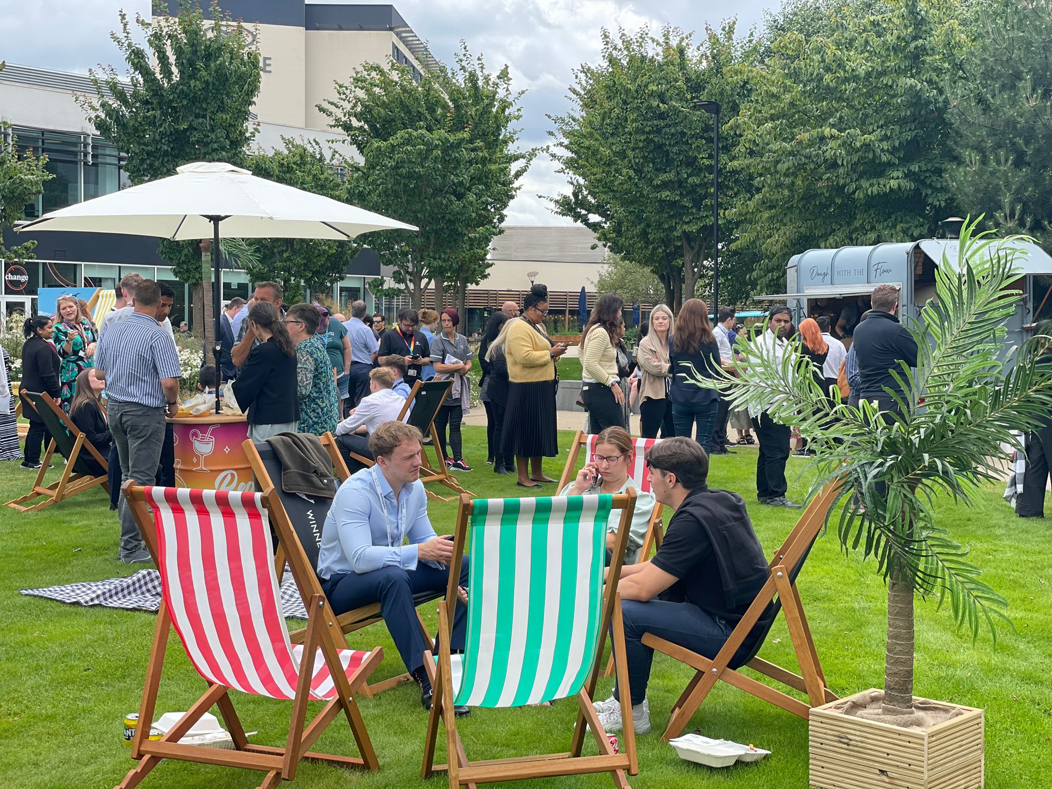 People are socialising outdoors on a grassy area with striped deckchairs, a food van, and a parasol, surrounded by trees and modern buildings.
