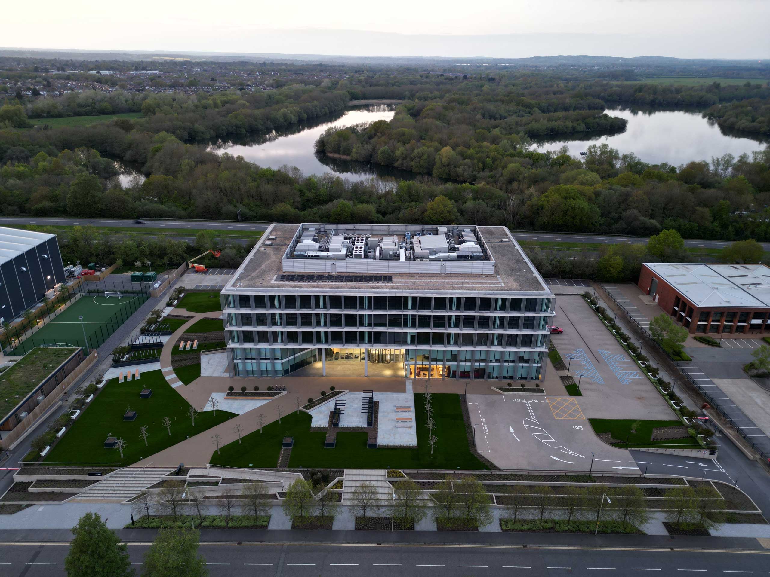 Aerial view of a modern, rectangular building with a flat roof, surrounded by green lawns, parking spaces, and a river in the background.