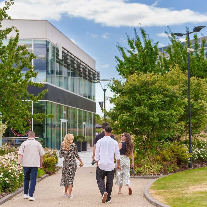 Five people walk along a path surrounded by greenery near a modern building with large windows on a partly cloudy day.